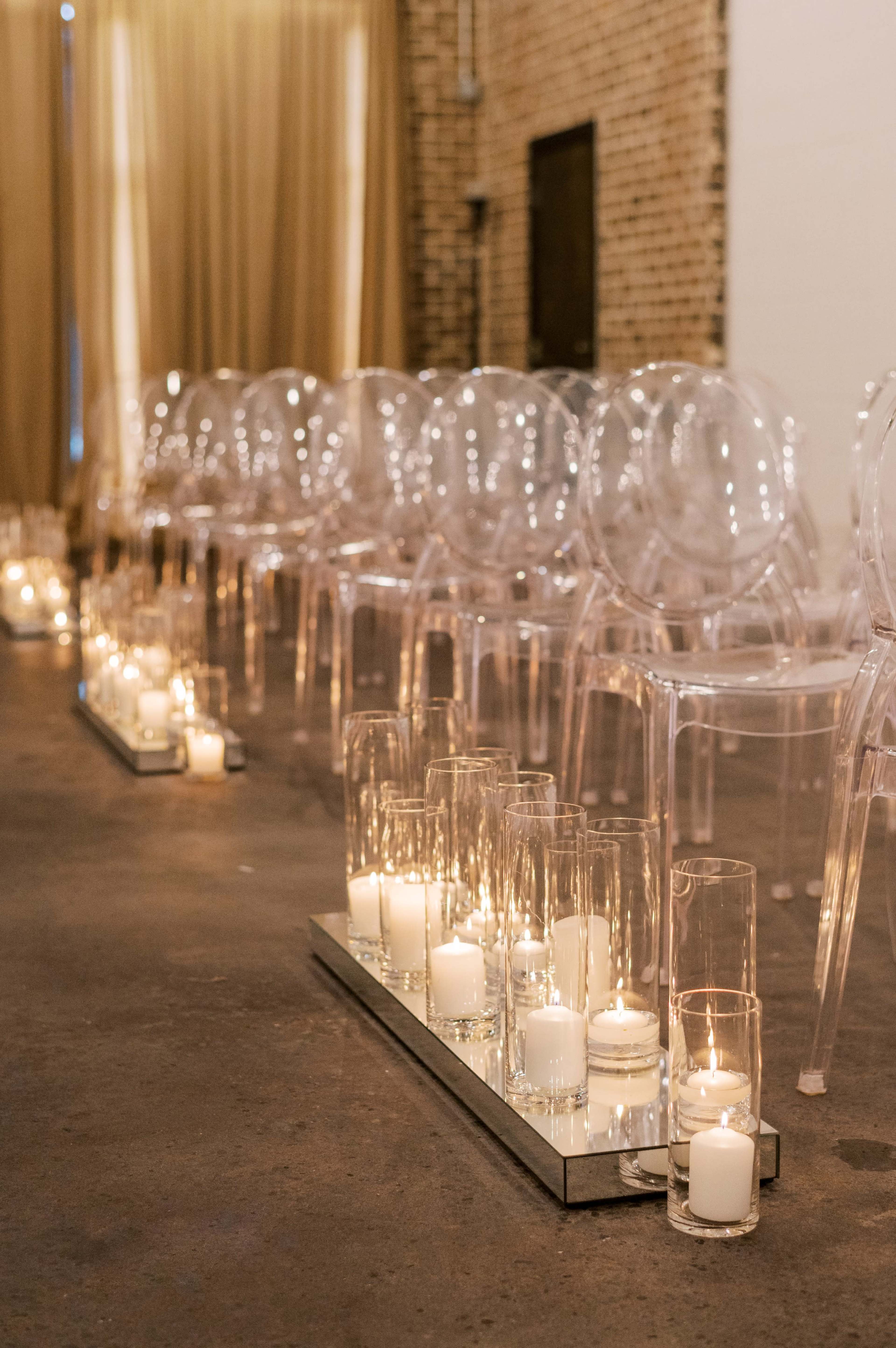 The image shows a row of clear, modern chairs beside a path lined with glass candle holders, creating a decorative setup for a ceremony.