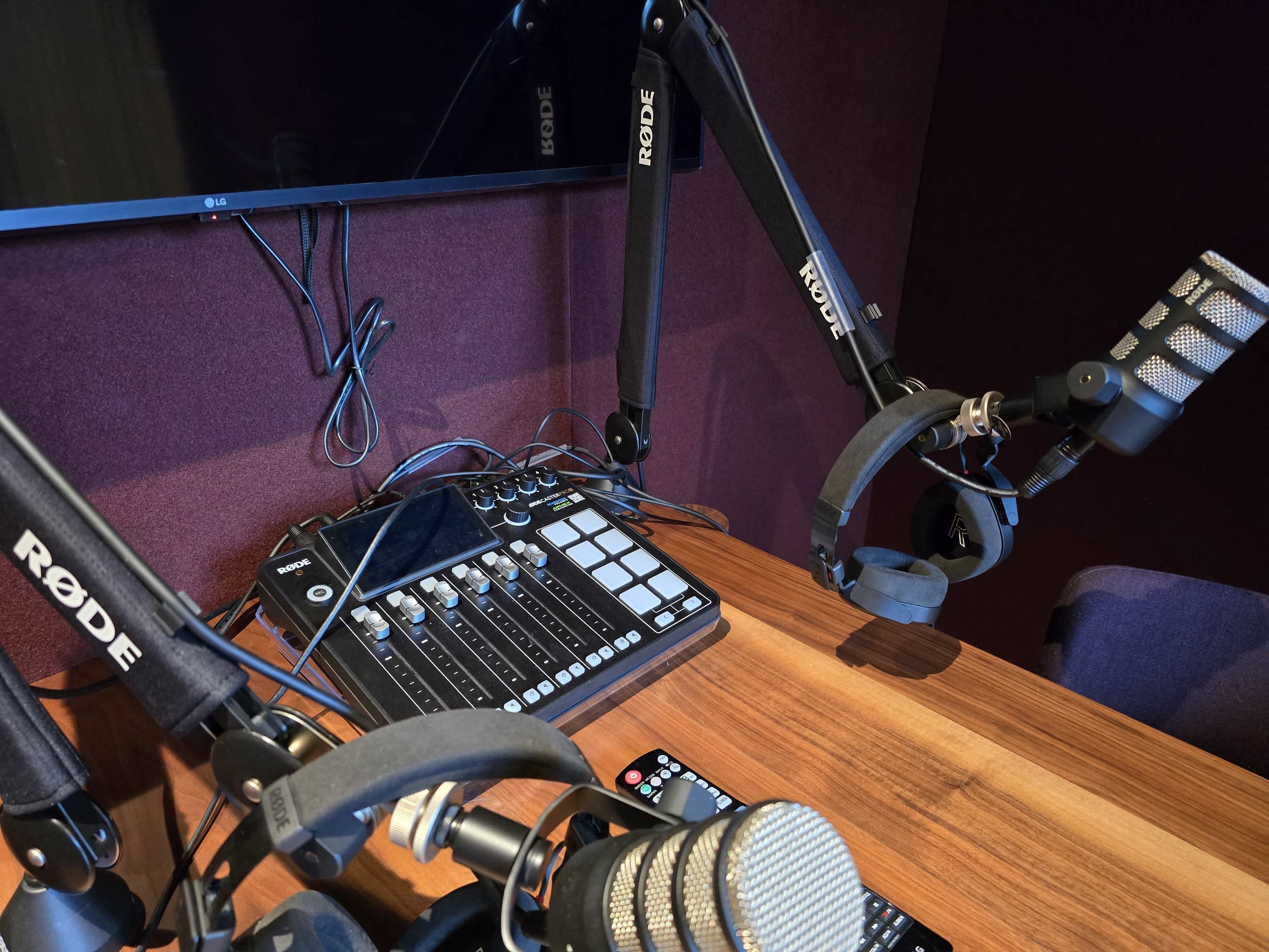 The image shows a recording studio setup featuring two microphones on adjustable arms, an audio mixer, and a television screen mounted on the wall.