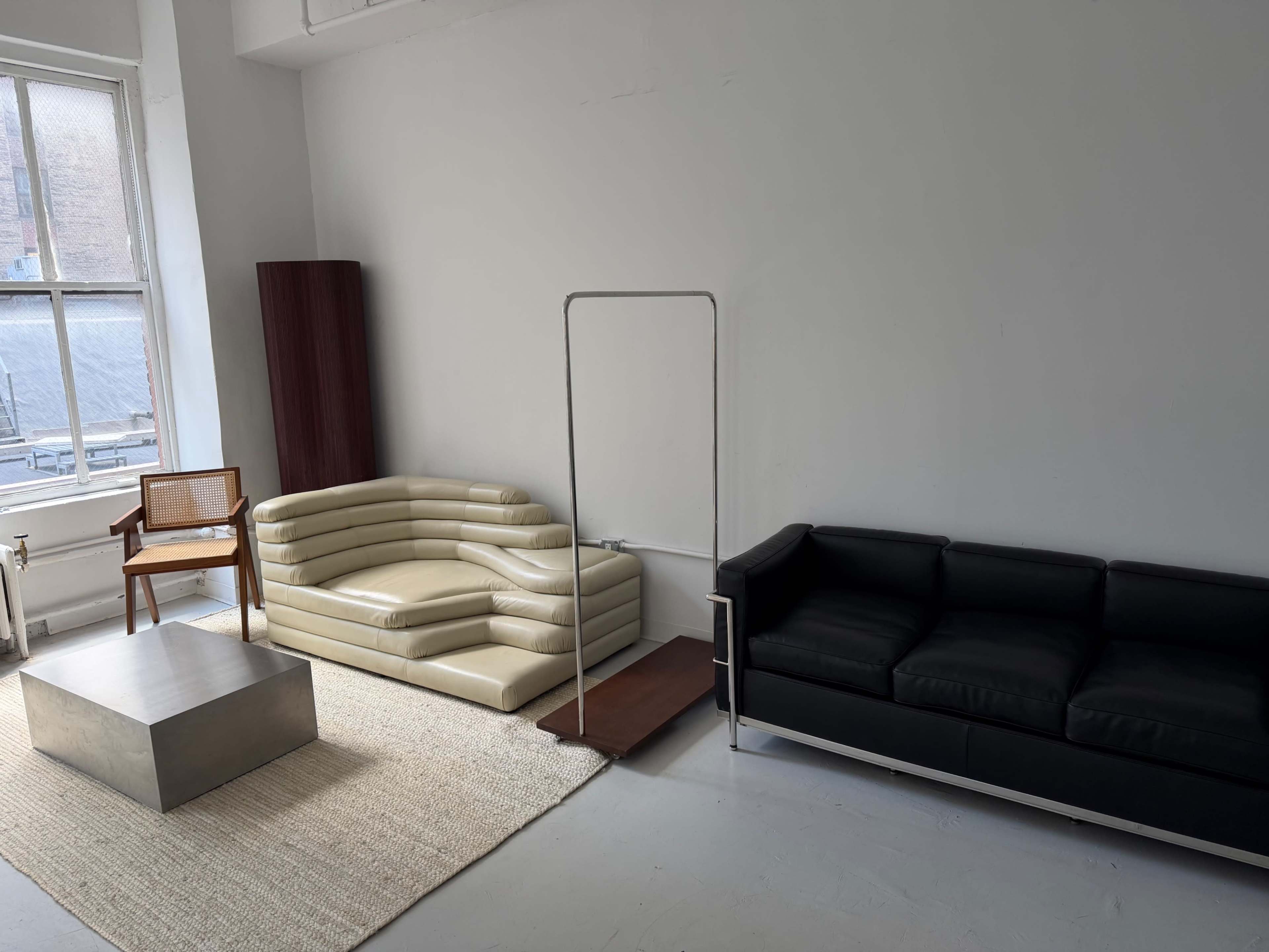 The image shows a minimalist living space featuring a beige curved sofa, a black leather couch, a wooden chair, a metal coffee table, and a wooden screen all set against a white wall with a window.