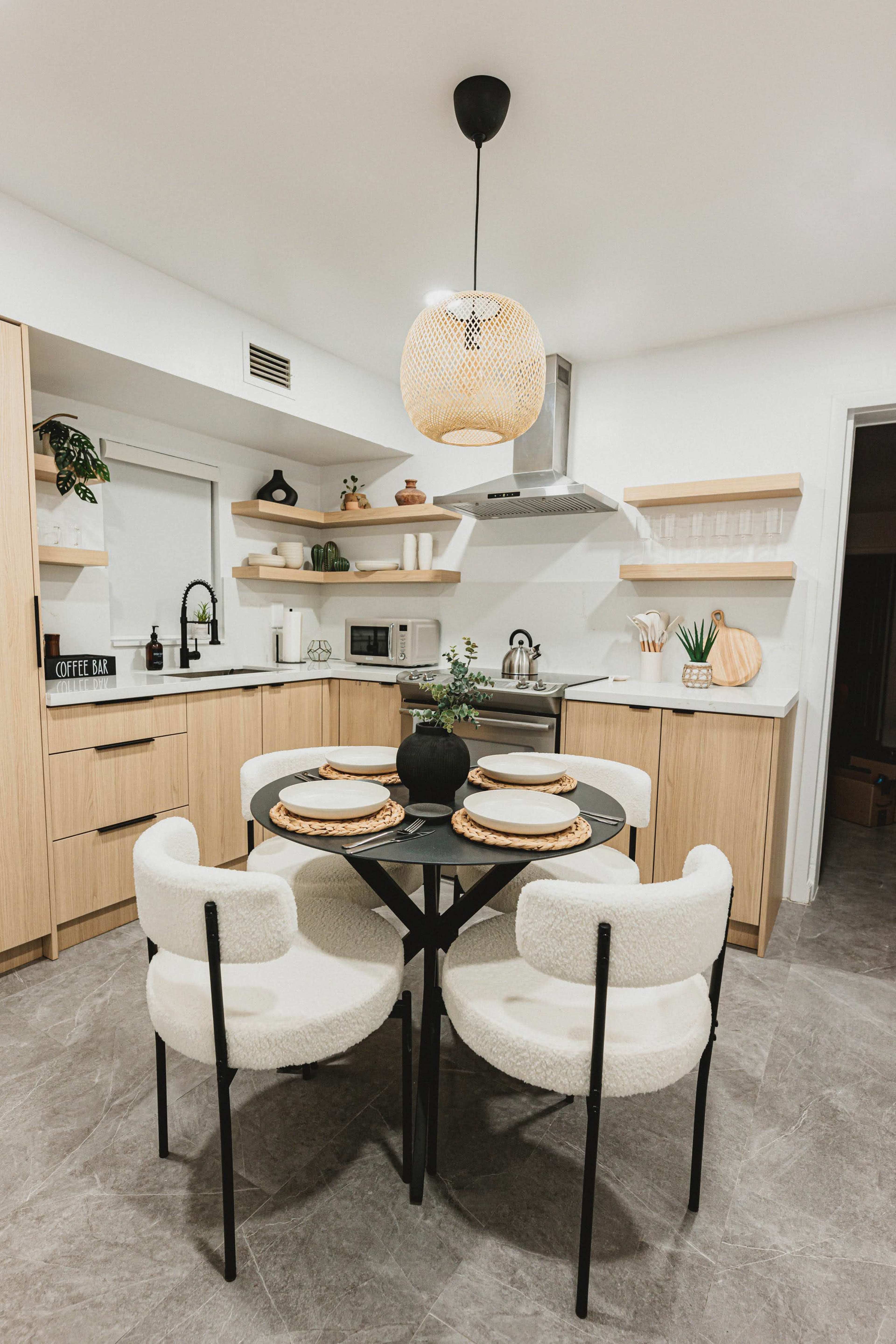The image shows a modern kitchen featuring a round black table with white, upholstered chairs, surrounded by wooden cabinetry and shelves, and a pendant light hanging from the ceiling.