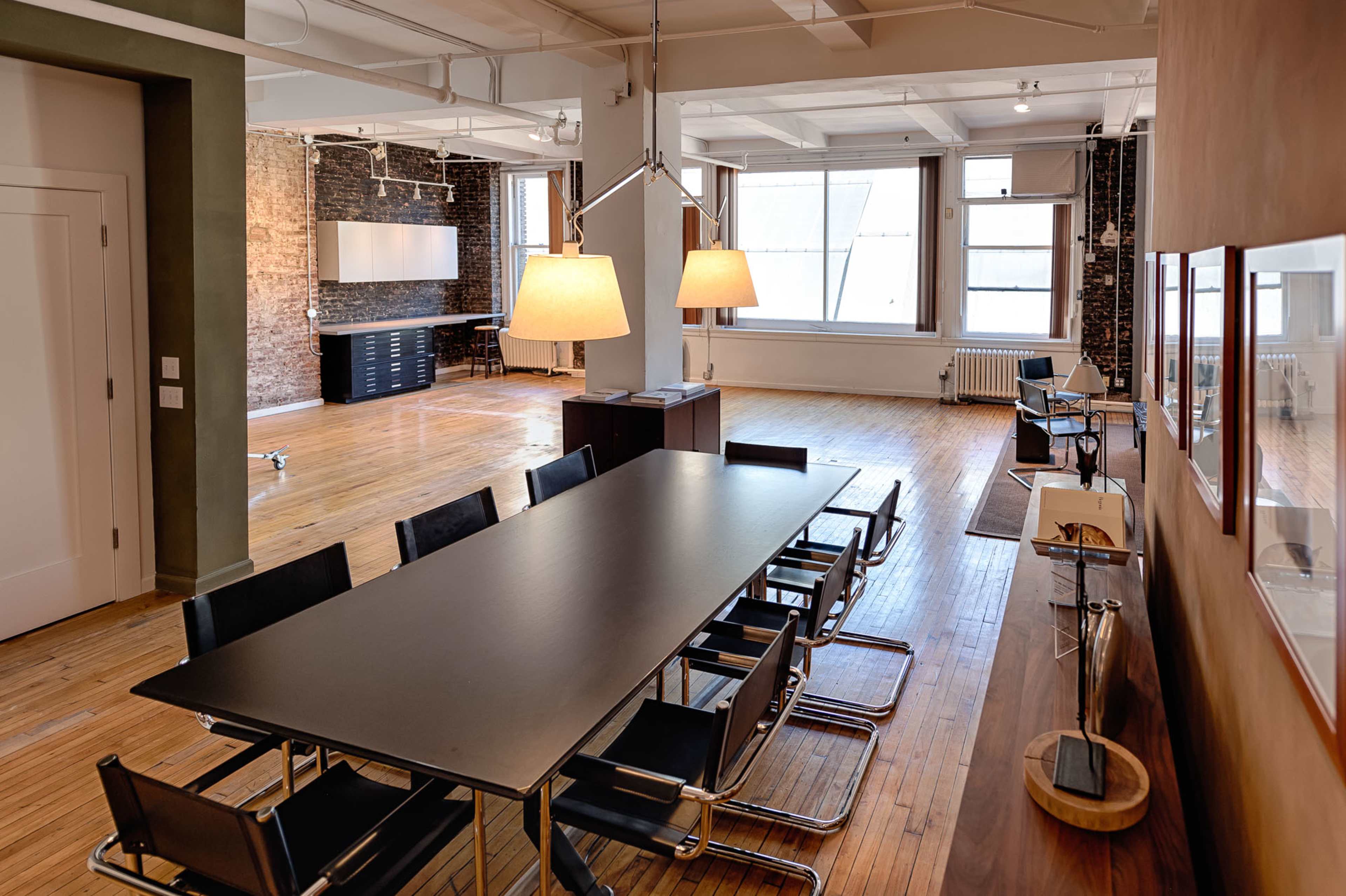 A modern open office space featuring a long black table surrounded by black chairs, with large windows allowing natural light to fill the room and exposed brick walls adding texture.