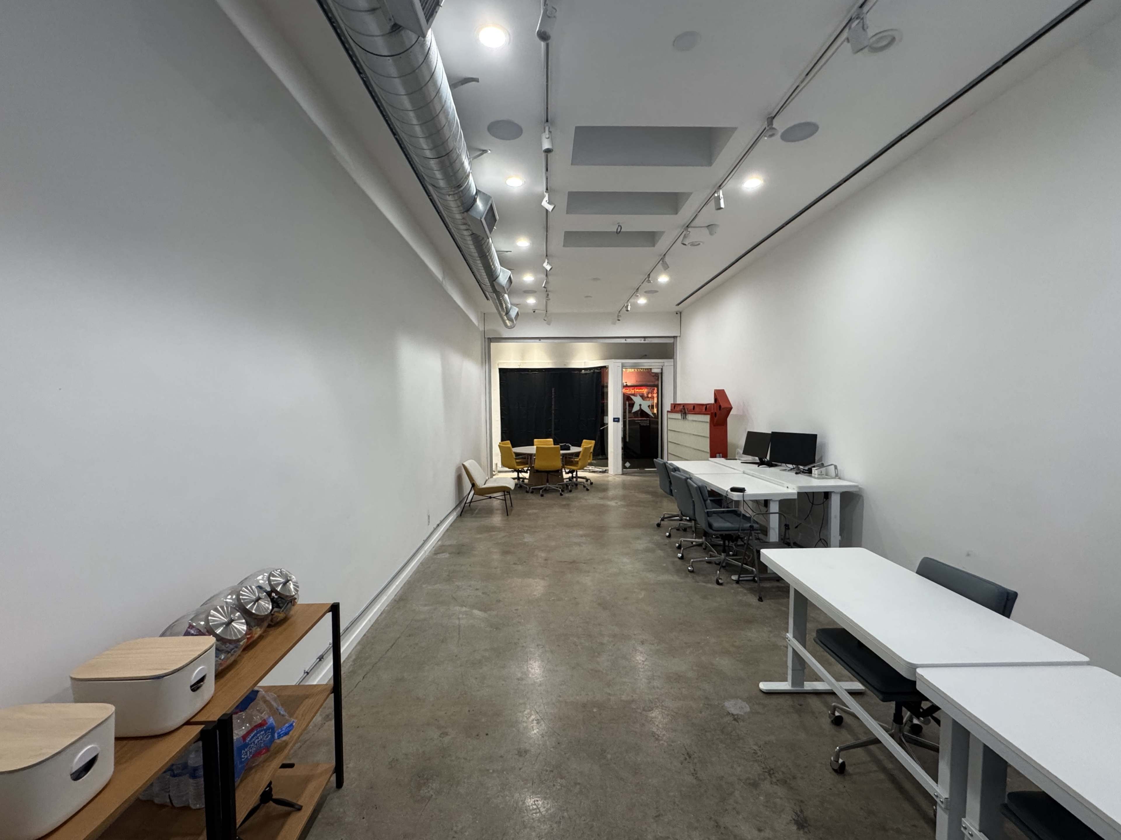 The image shows a long, narrow office hallway featuring desks, chairs, and shelves along the walls, with a black curtain at the far end.