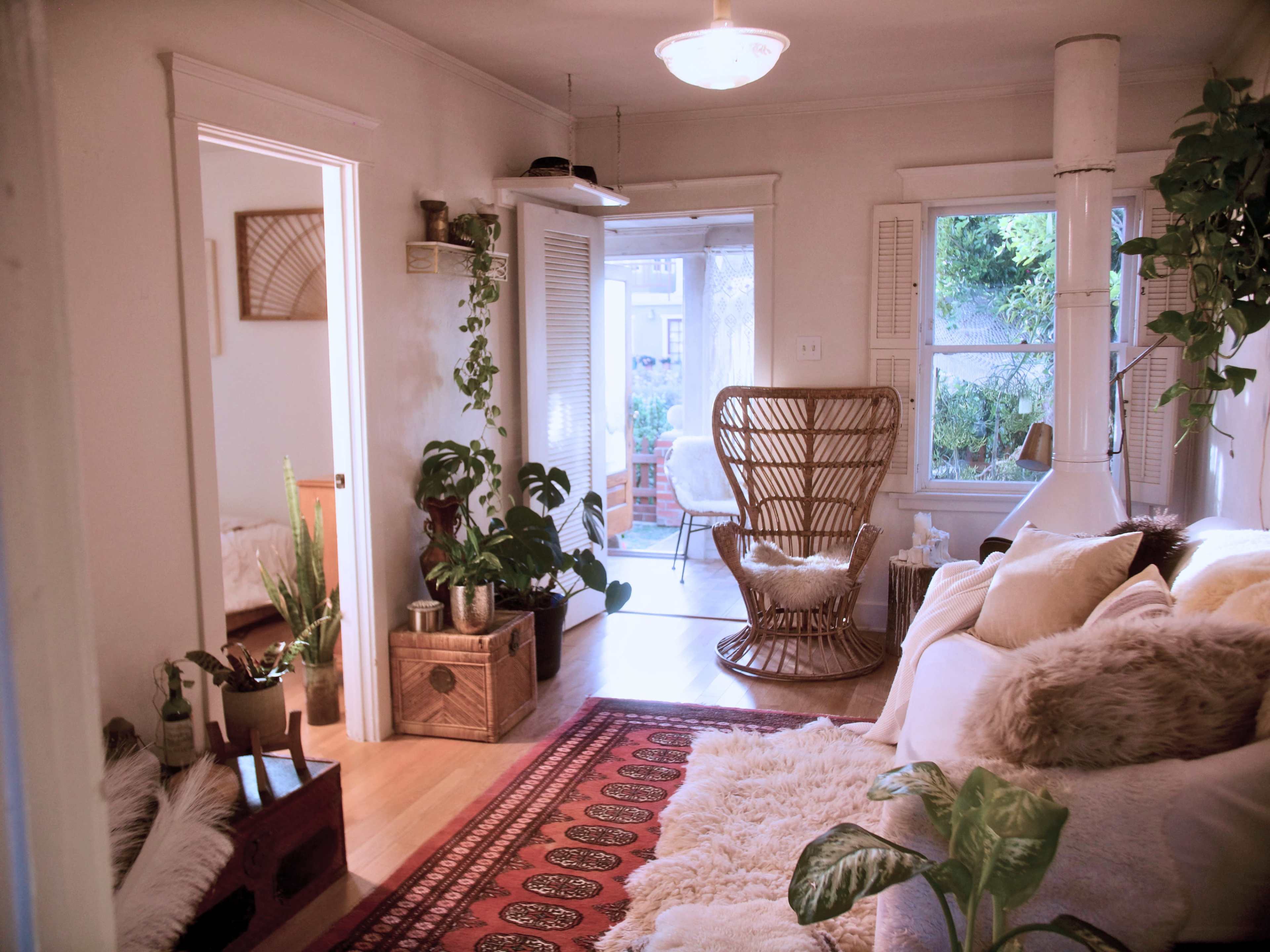 The image shows a cozy living room decorated with plants, a large woven chair, a sofa with blankets, and natural light coming through windows.