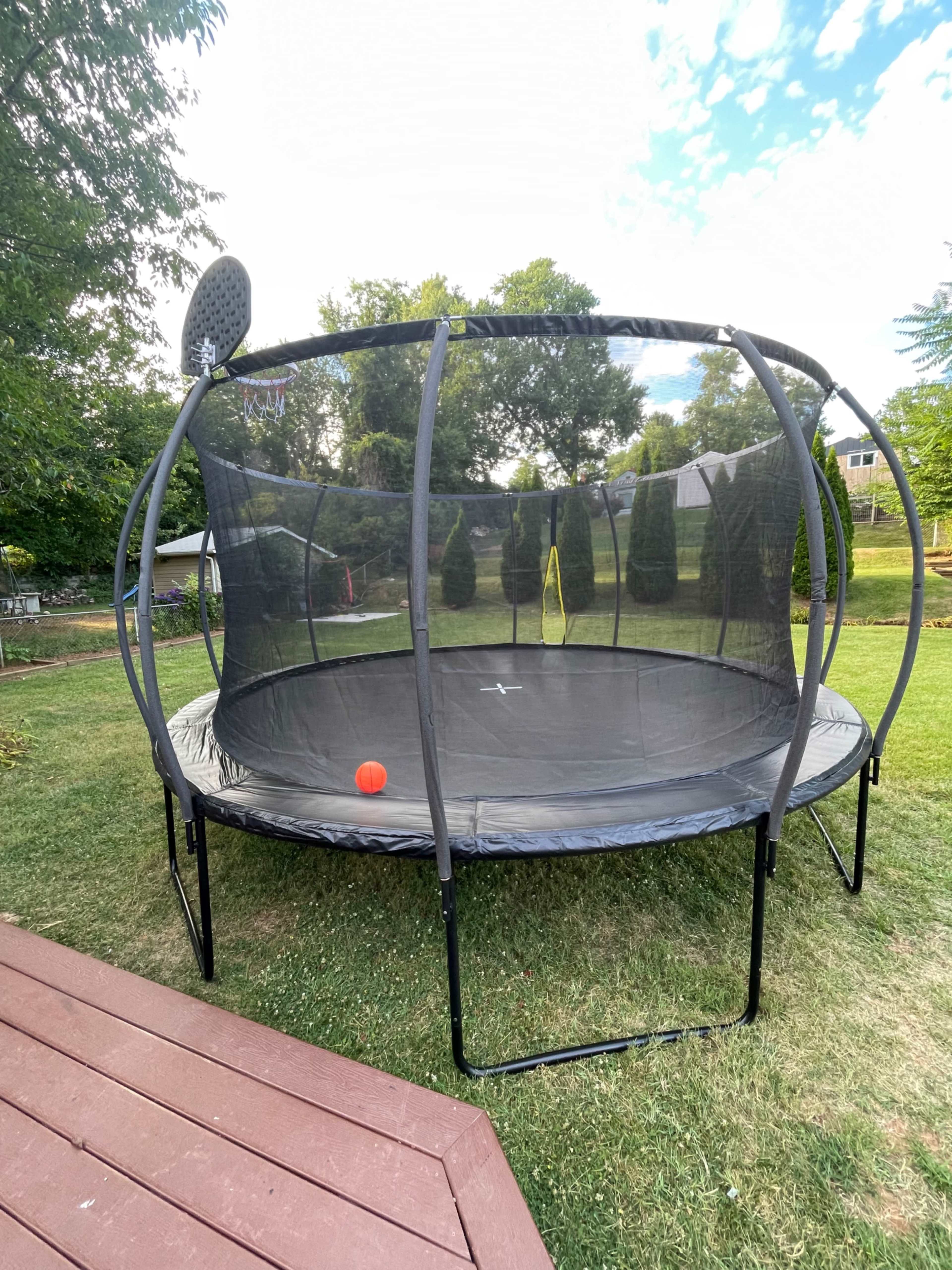 A round trampoline with a safety net and a basketball hoop is set up on a grassy lawn near a wooden deck.