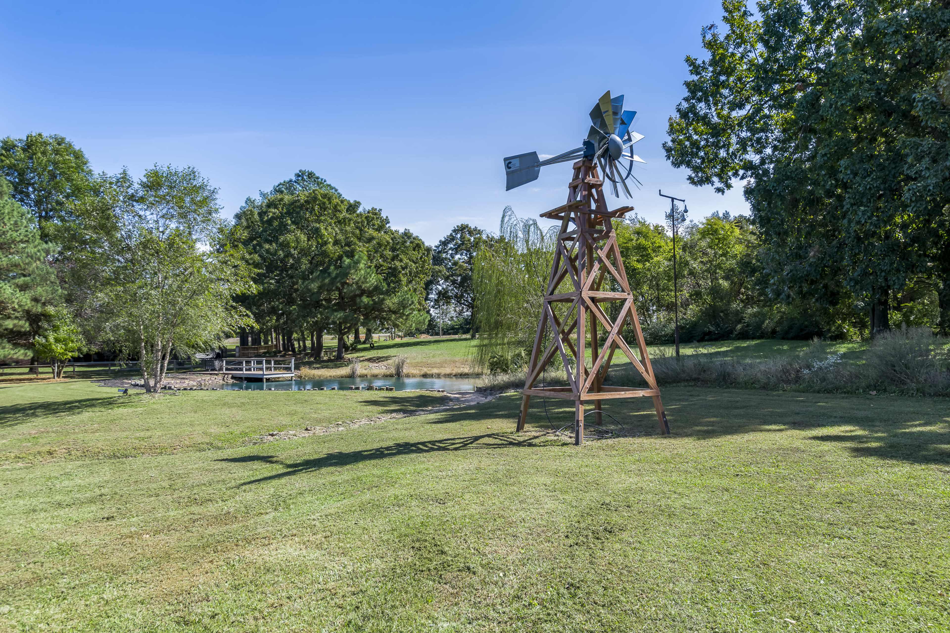 A wooden windmill stands in a grassy field near a pond, surrounded by trees and a wooden dock.