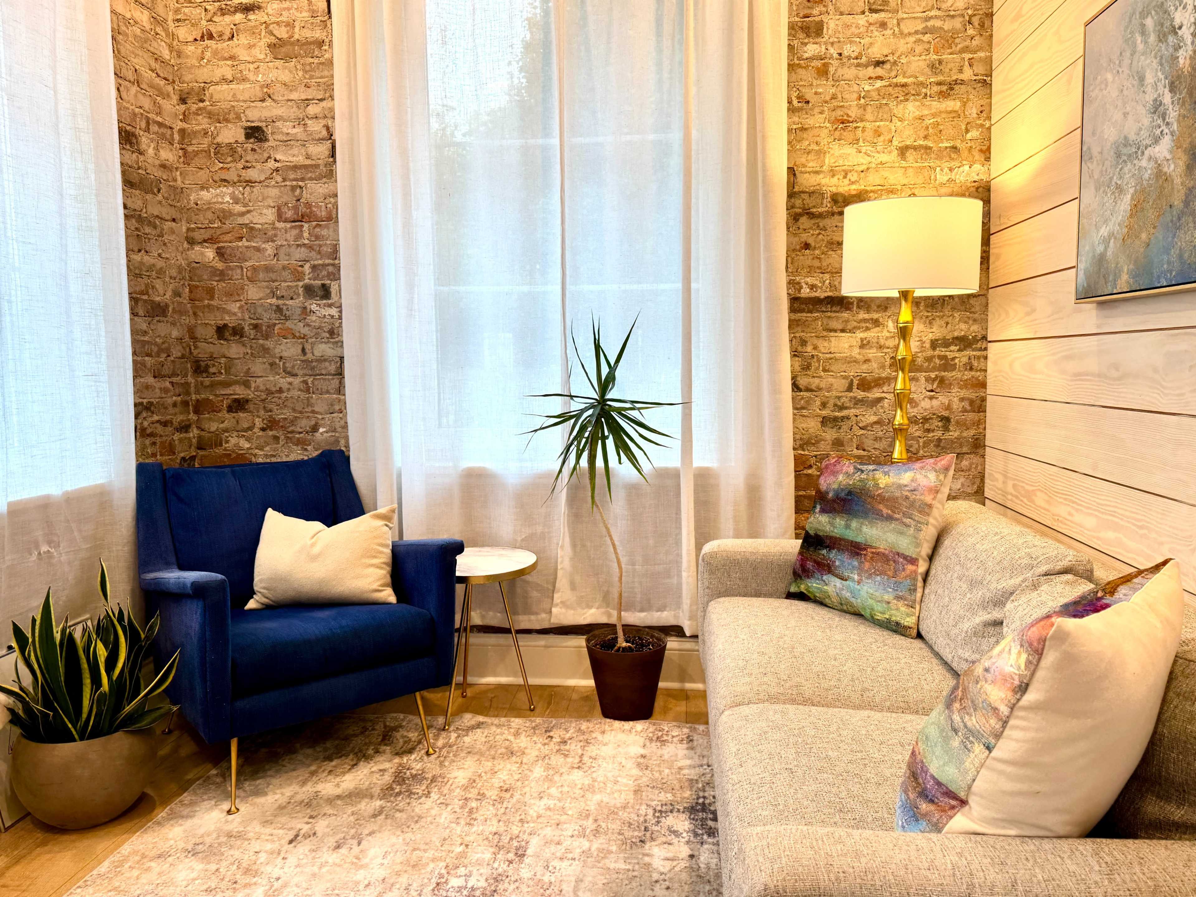 The image shows a cozy living space with a blue armchair and a light gray sofa, surrounded by textured brick walls and a tall indoor plant.