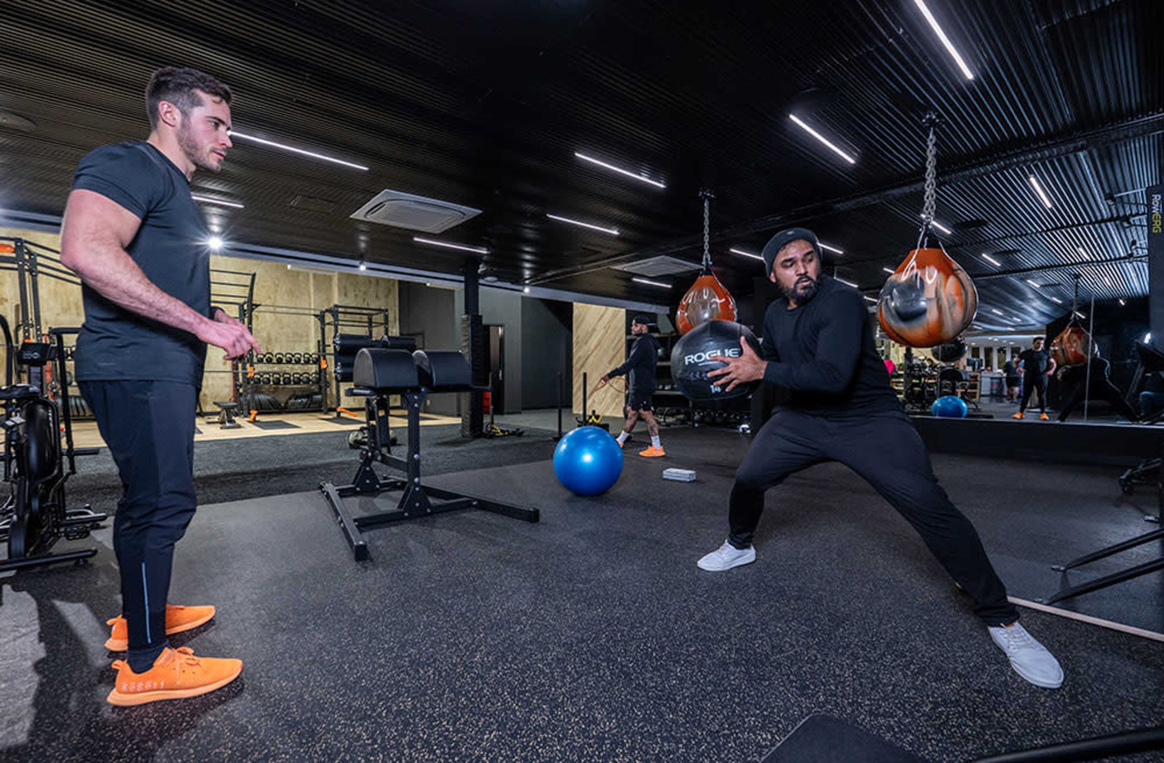 Two men are engaged in a workout in a gym, with one performing a medicine ball exercise while the other observes.