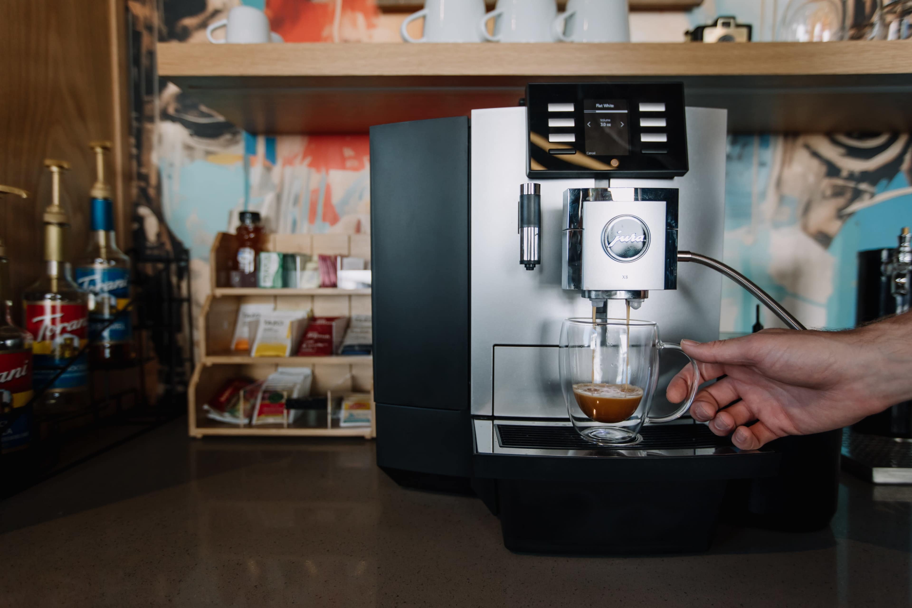 A person pours espresso from a silver coffee machine into a transparent glass cup on a countertop.