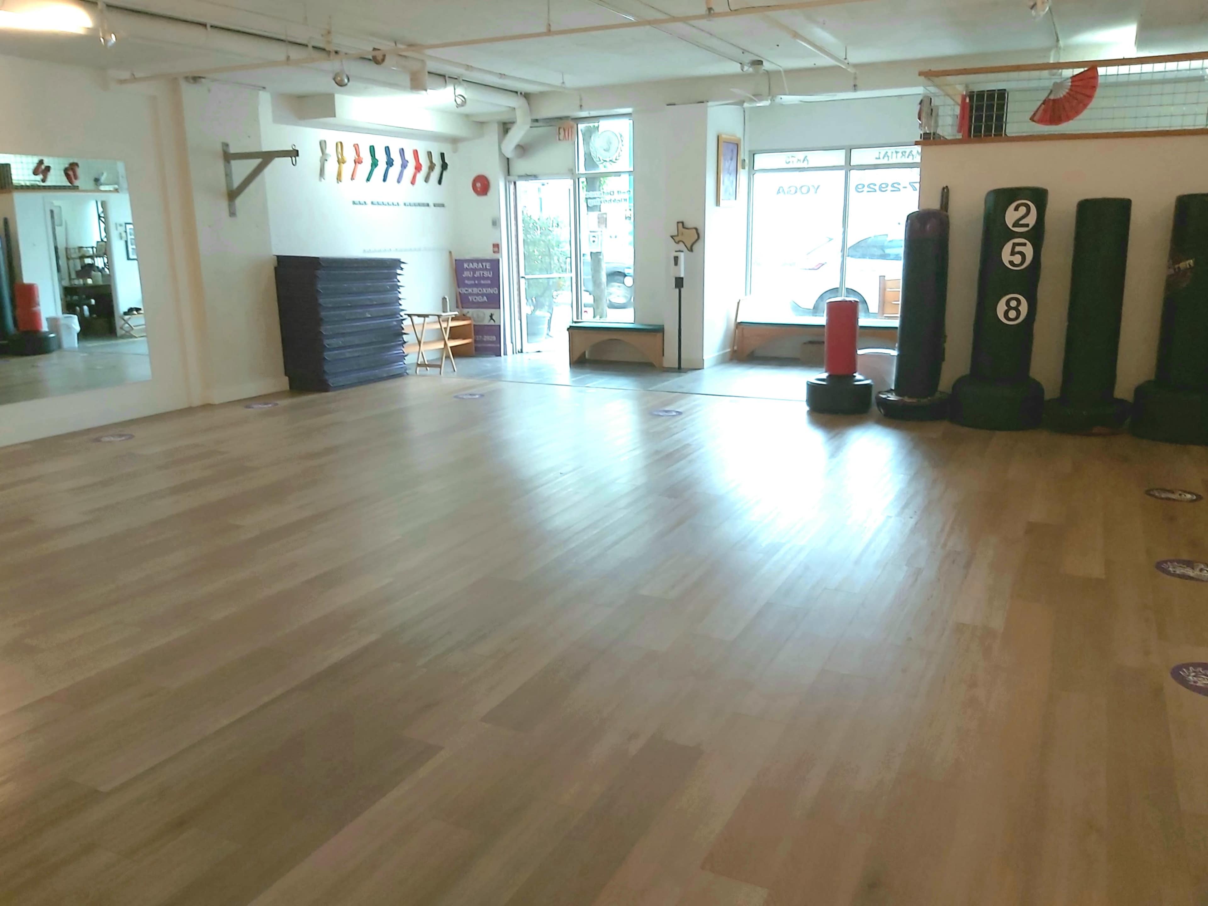 The image shows a spacious martial arts studio with wooden flooring, training equipment along the walls, and a large window allowing natural light to enter.