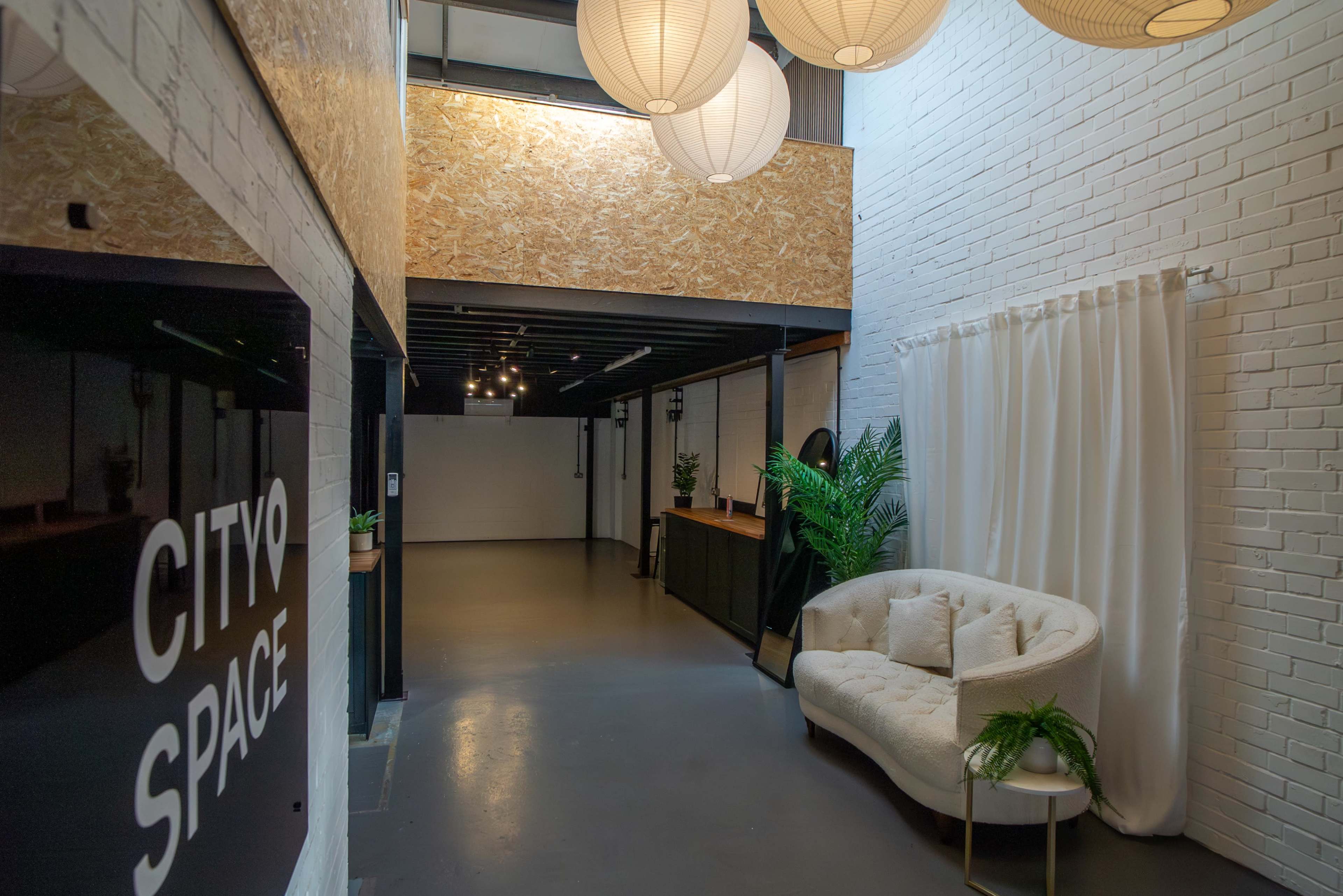 The image shows a modern, well-lit interior space featuring a couch, decorative plants, and pendant lights, with a sign reading "CITY SPACE" on the left.