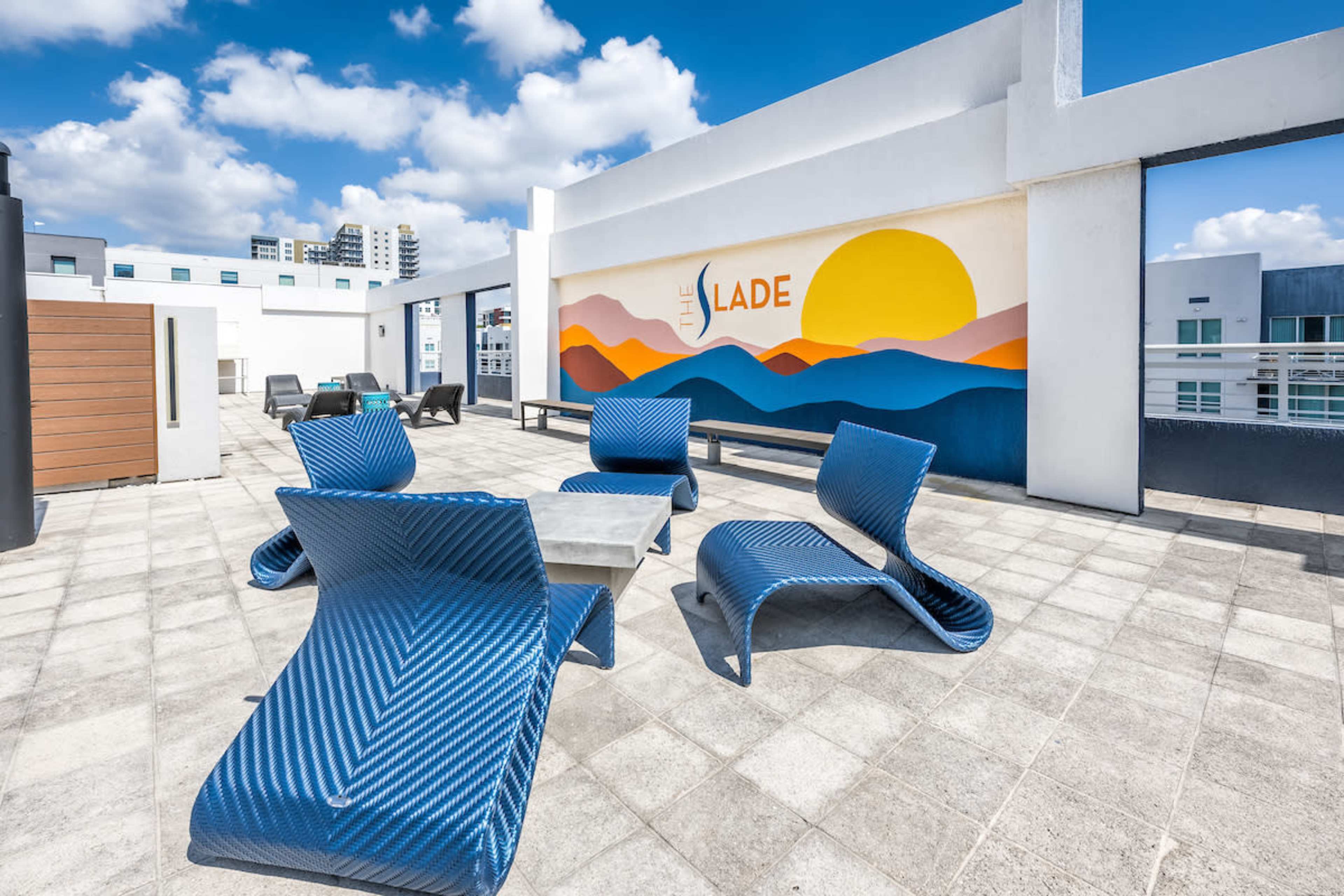 A rooftop terrace with blue lounge chairs, a table, and a colorful mural of mountains and a sun in the background.