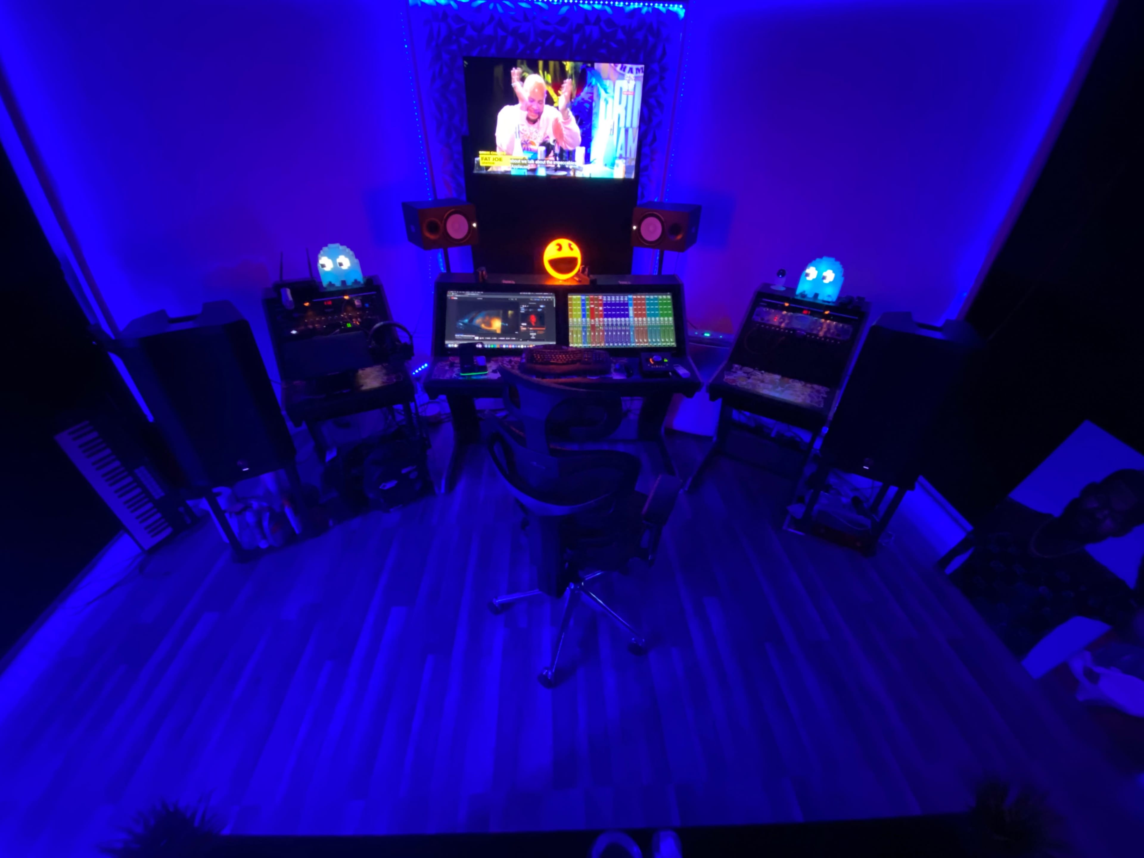 A modern music production studio illuminated with blue lights, featuring a central workstation with multiple screens, audio equipment, and speakers.
