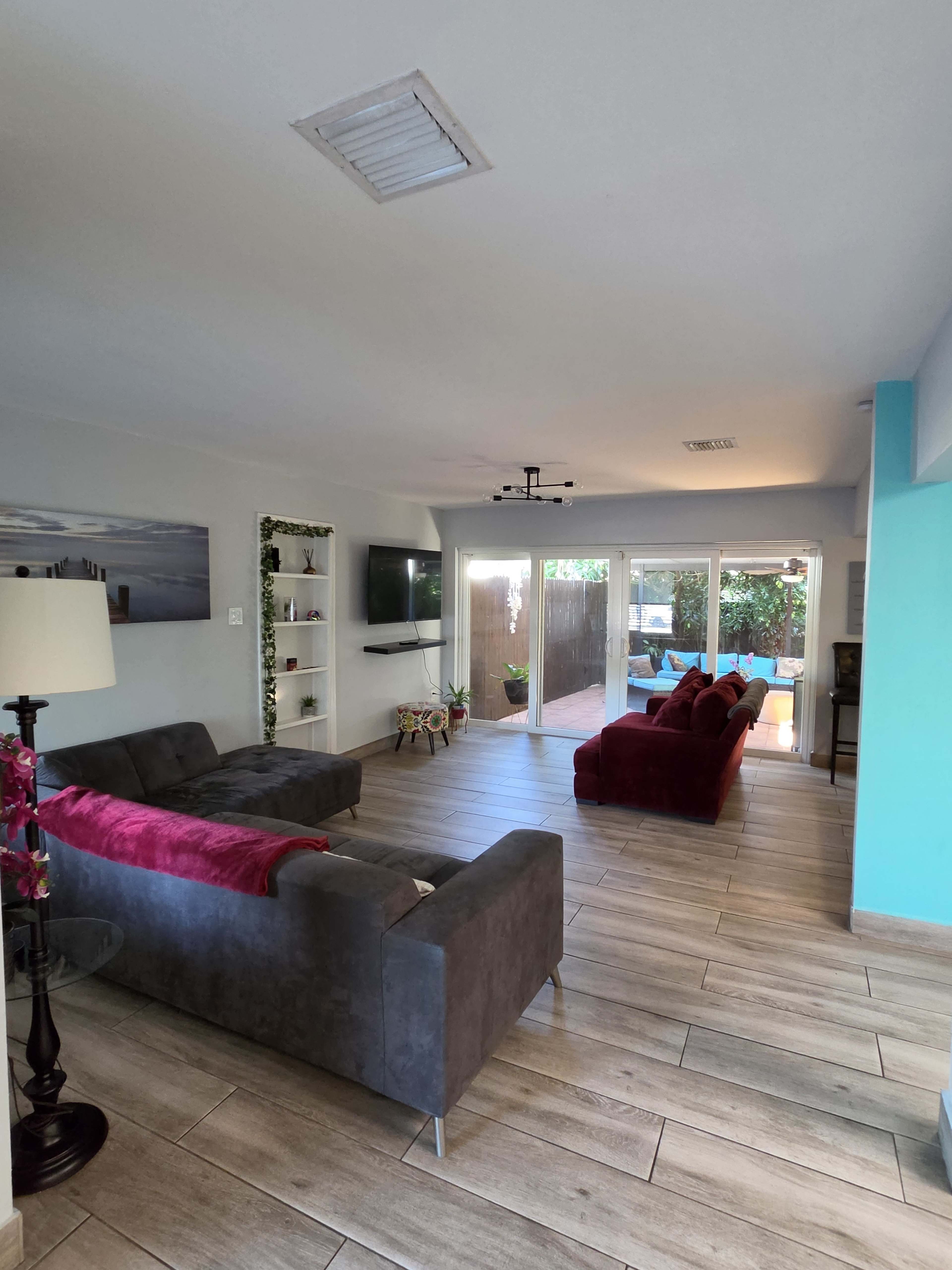 The image shows a modern living room with gray and maroon sofas, a television mounted on the wall, and large sliding glass doors leading to an outdoor area.