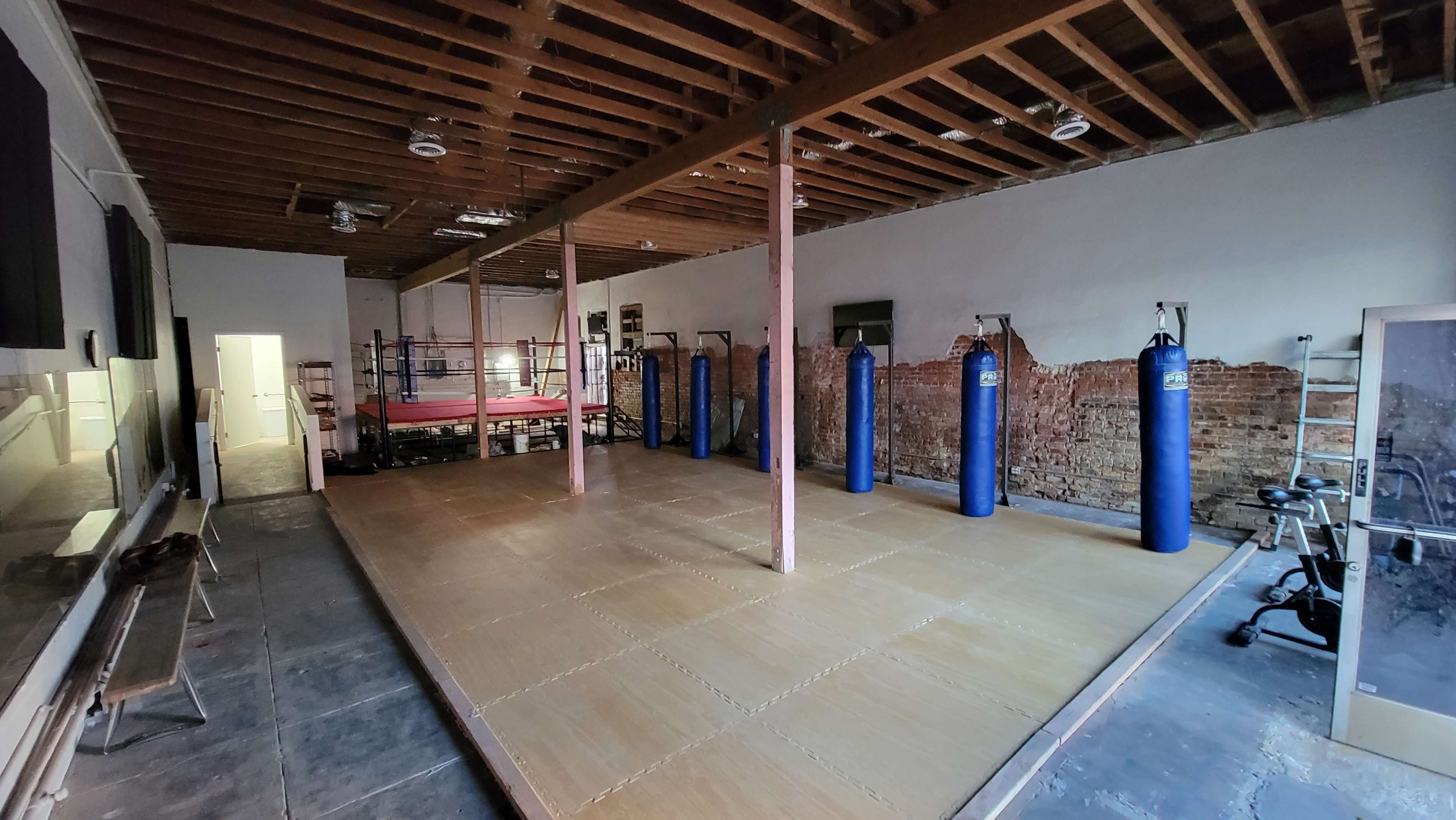 A training facility with a wooden mat area, several blue punching bags, and a boxing ring in the background.