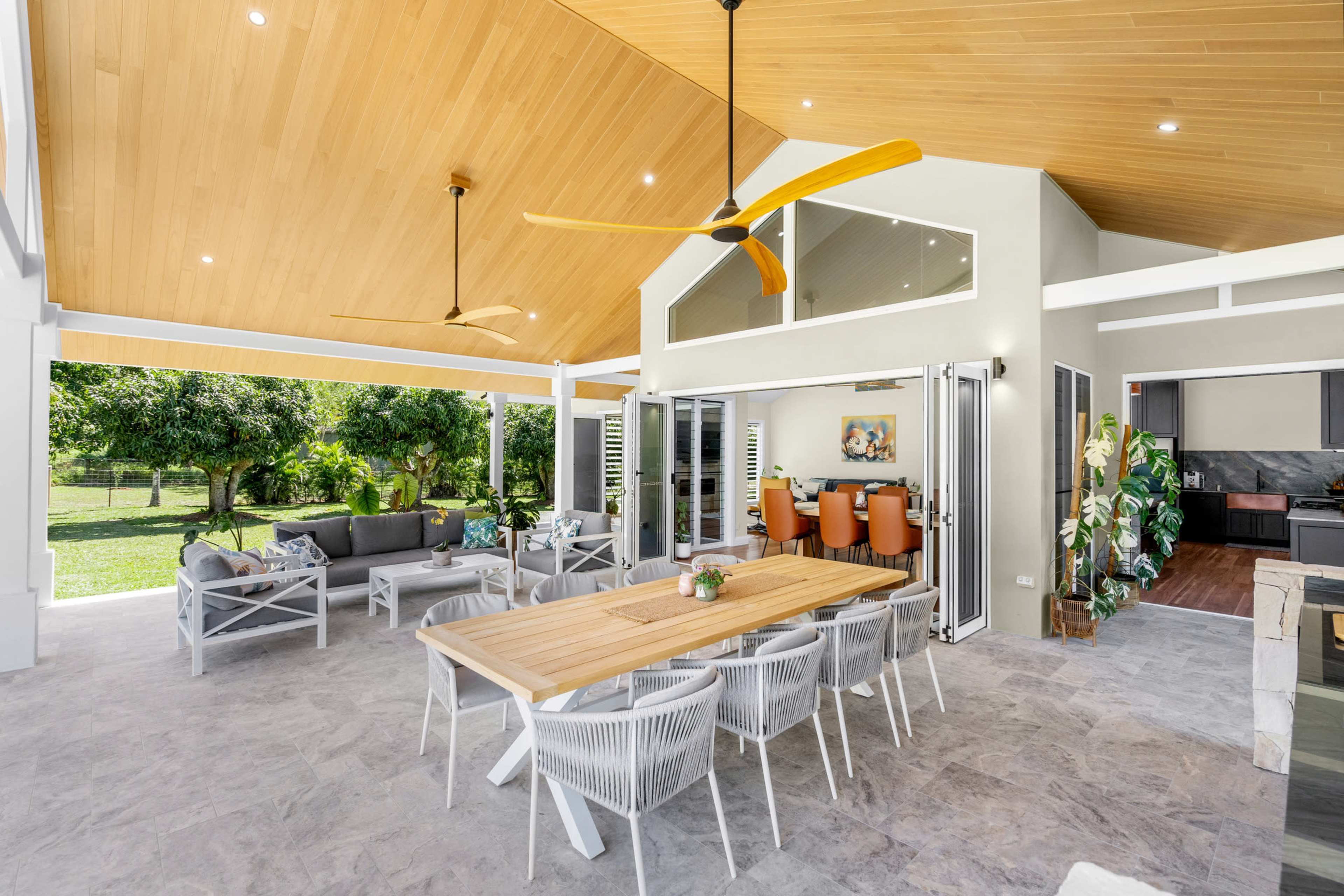 The image shows a modern outdoor living space with a spacious dining area, comfortable seating, and large windows opening to a lush garden.