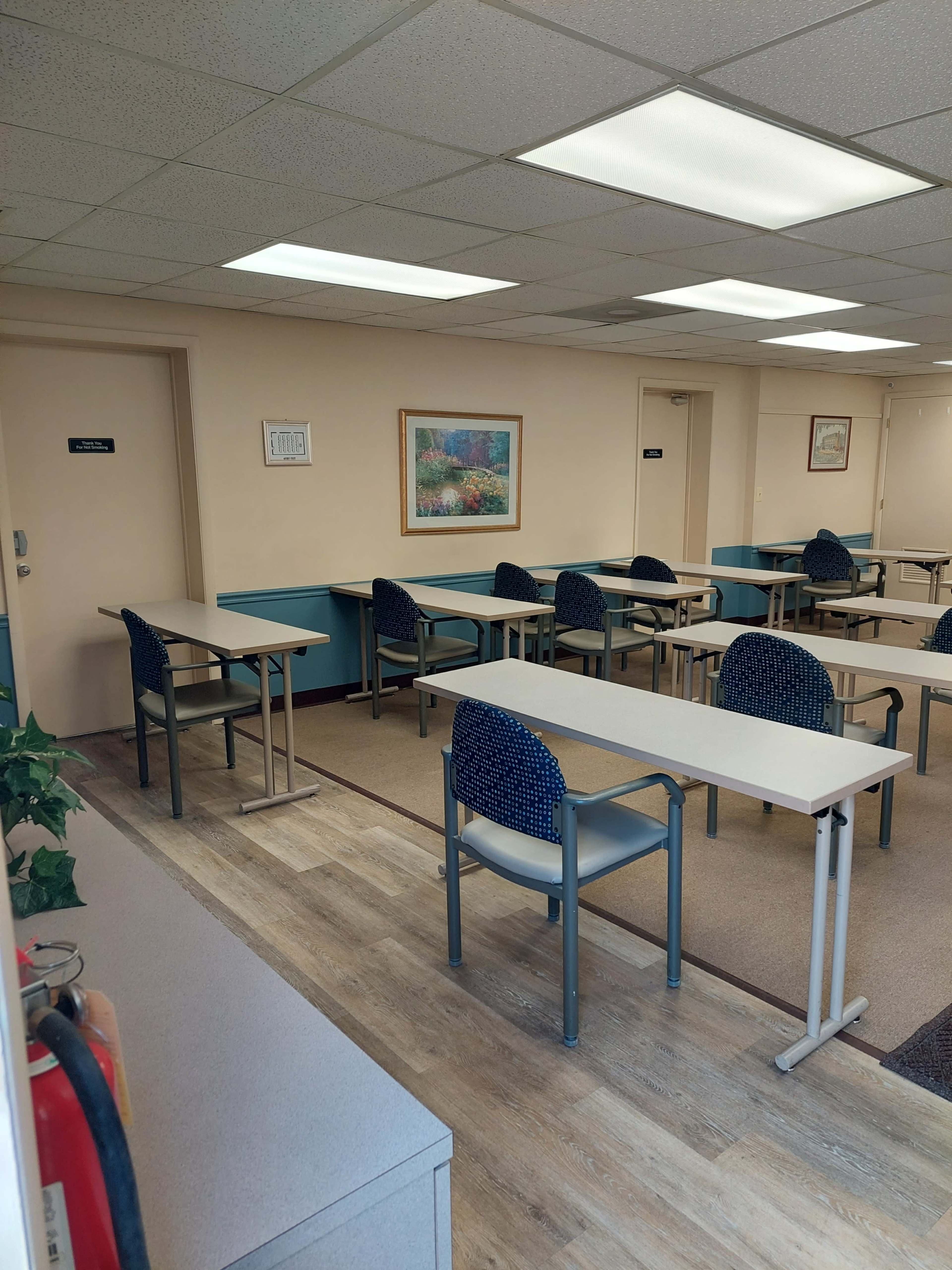 A classroom with several tables and chairs arranged in rows, along with a fire extinguisher and framed artwork on the walls.