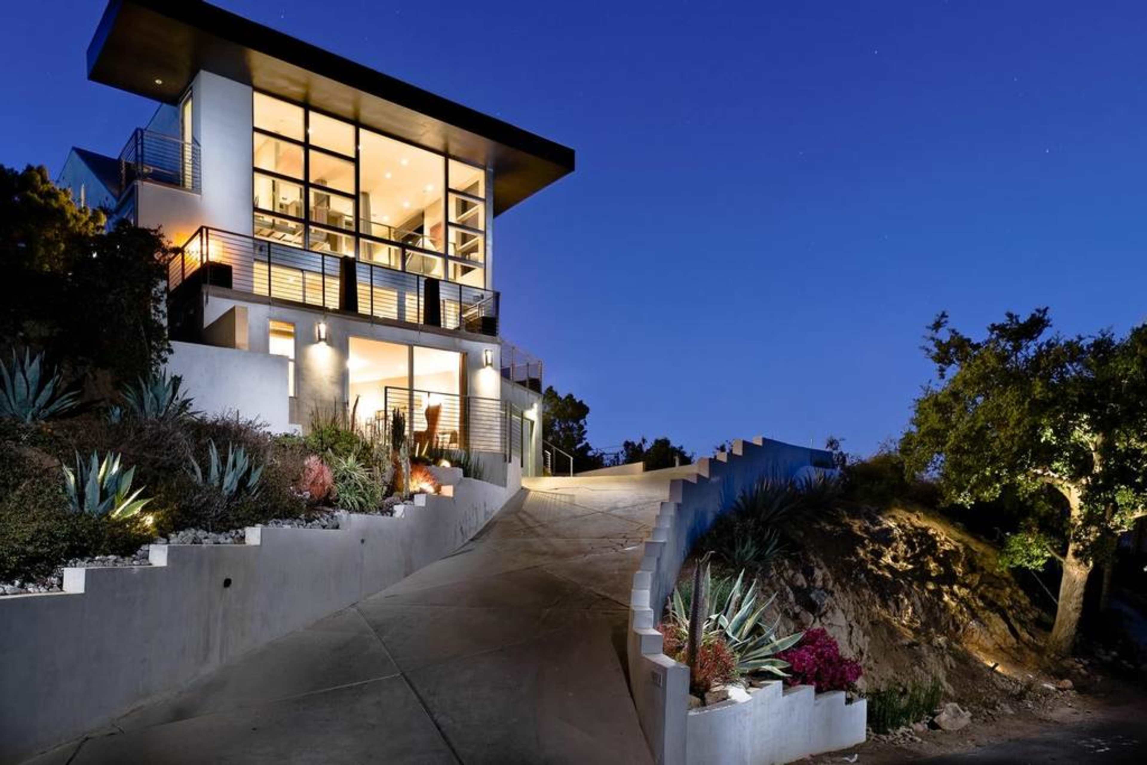 A modern house with large windows is situated on a sloped driveway, surrounded by landscaped greenery and lighting at dusk.