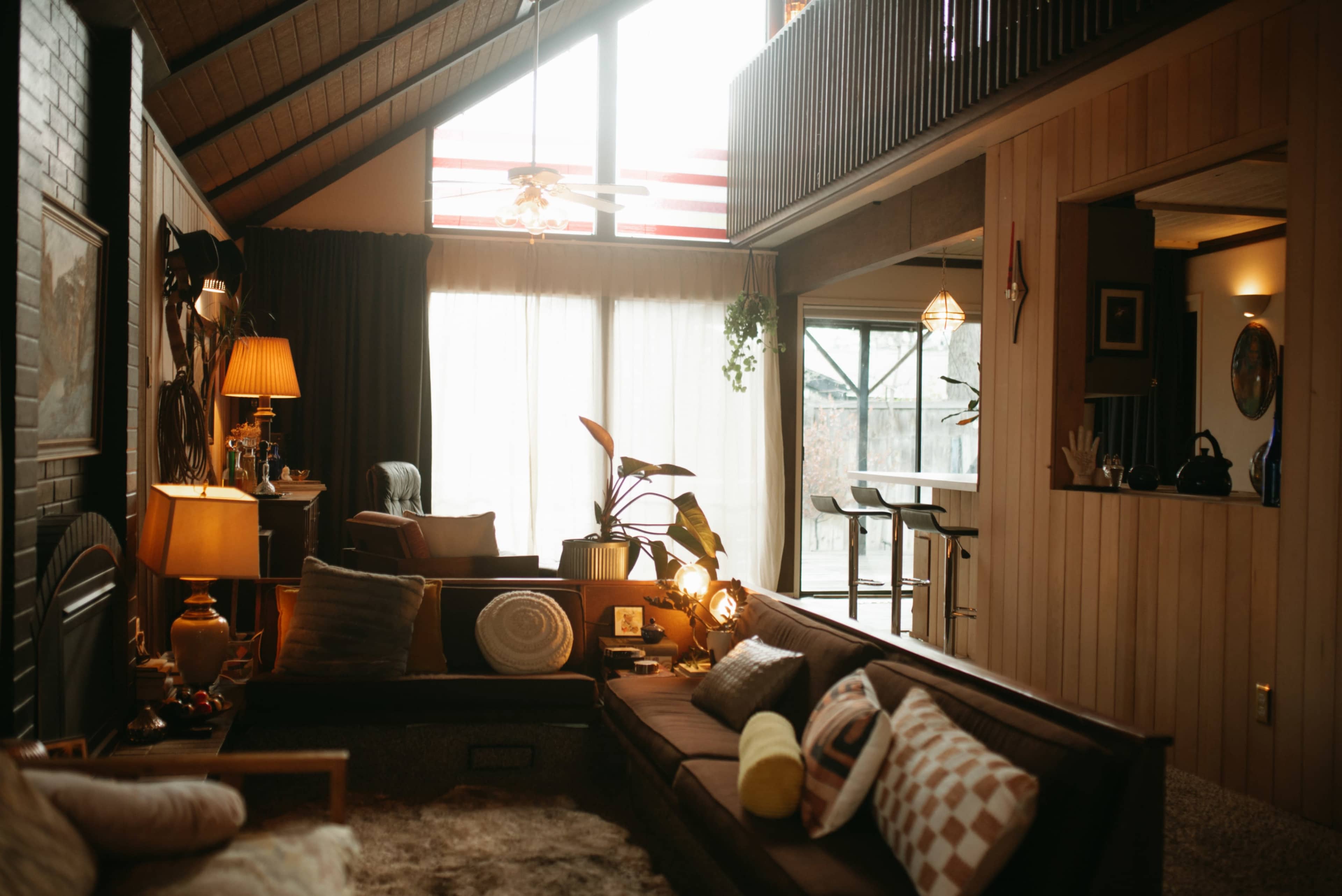 The image features a spacious, modern living room with large windows, a sectional sofa, and a mix of decorative lamps and plants.