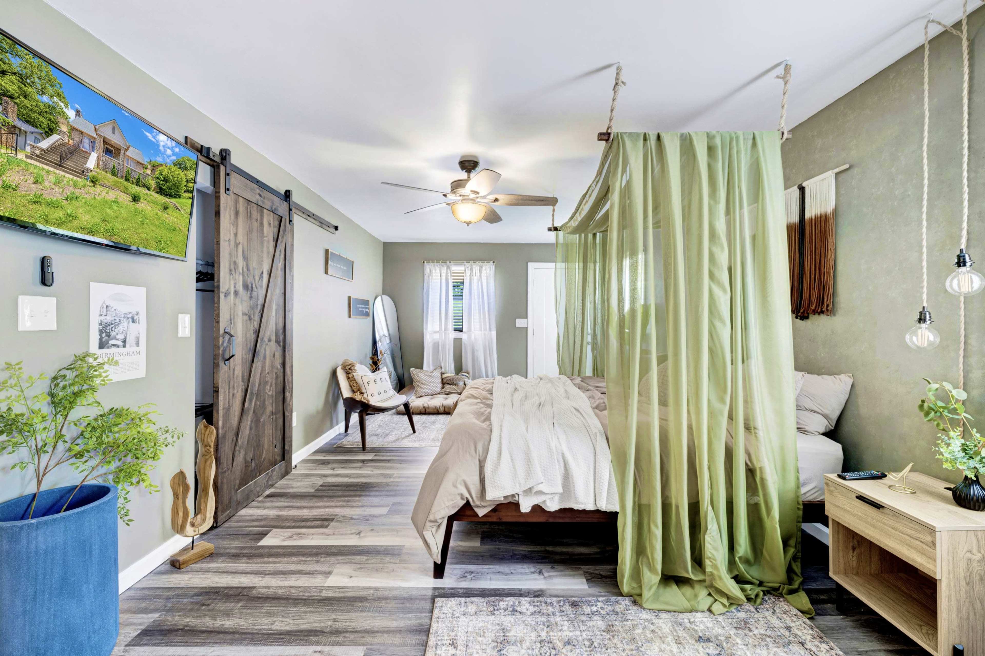The room features a bed with green sheer curtains, a television mounted on the wall, and a mix of wooden and modern furniture accents.