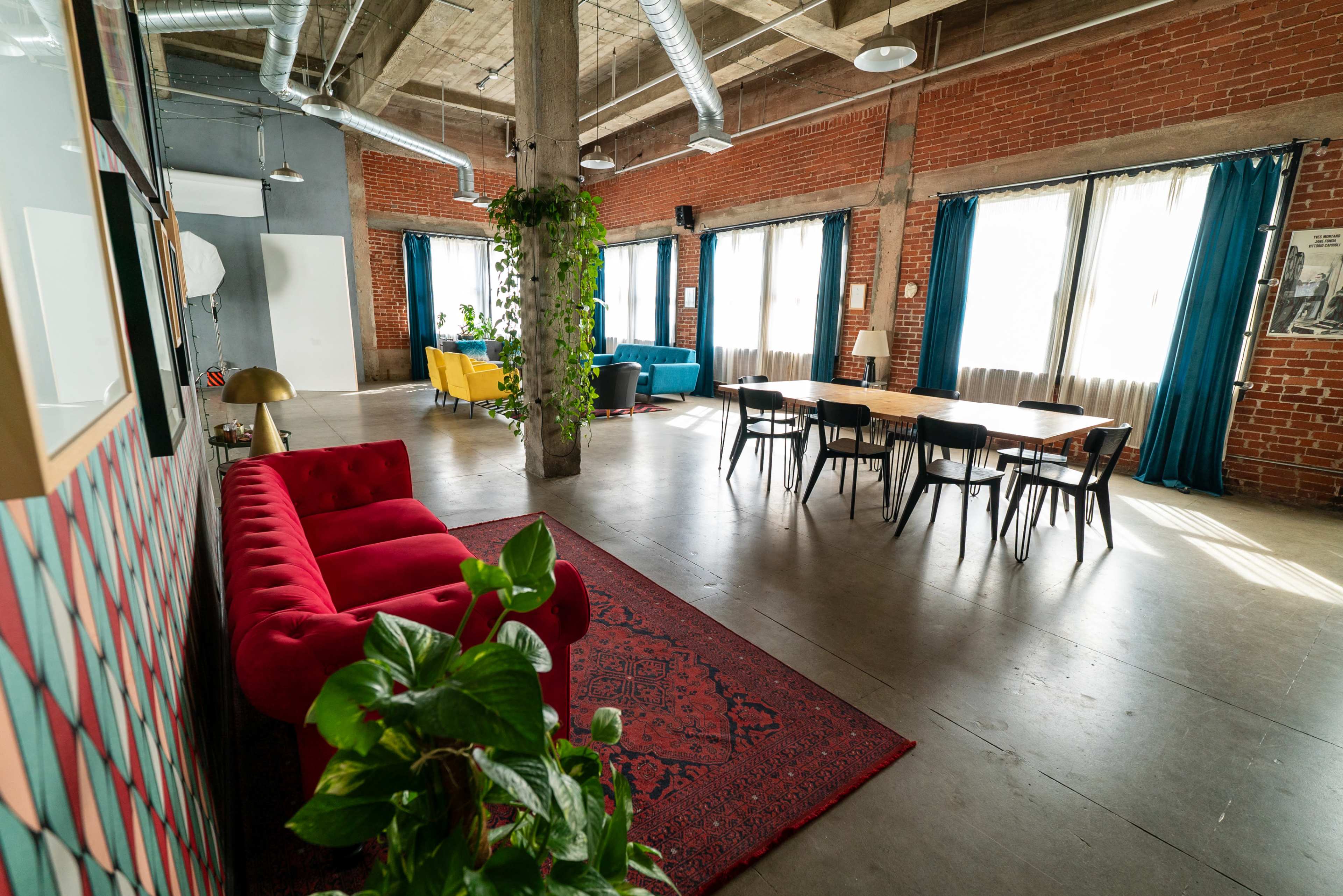The image shows a spacious, industrial-style room featuring red couches, a wooden dining table, and large windows with blue and green curtains.