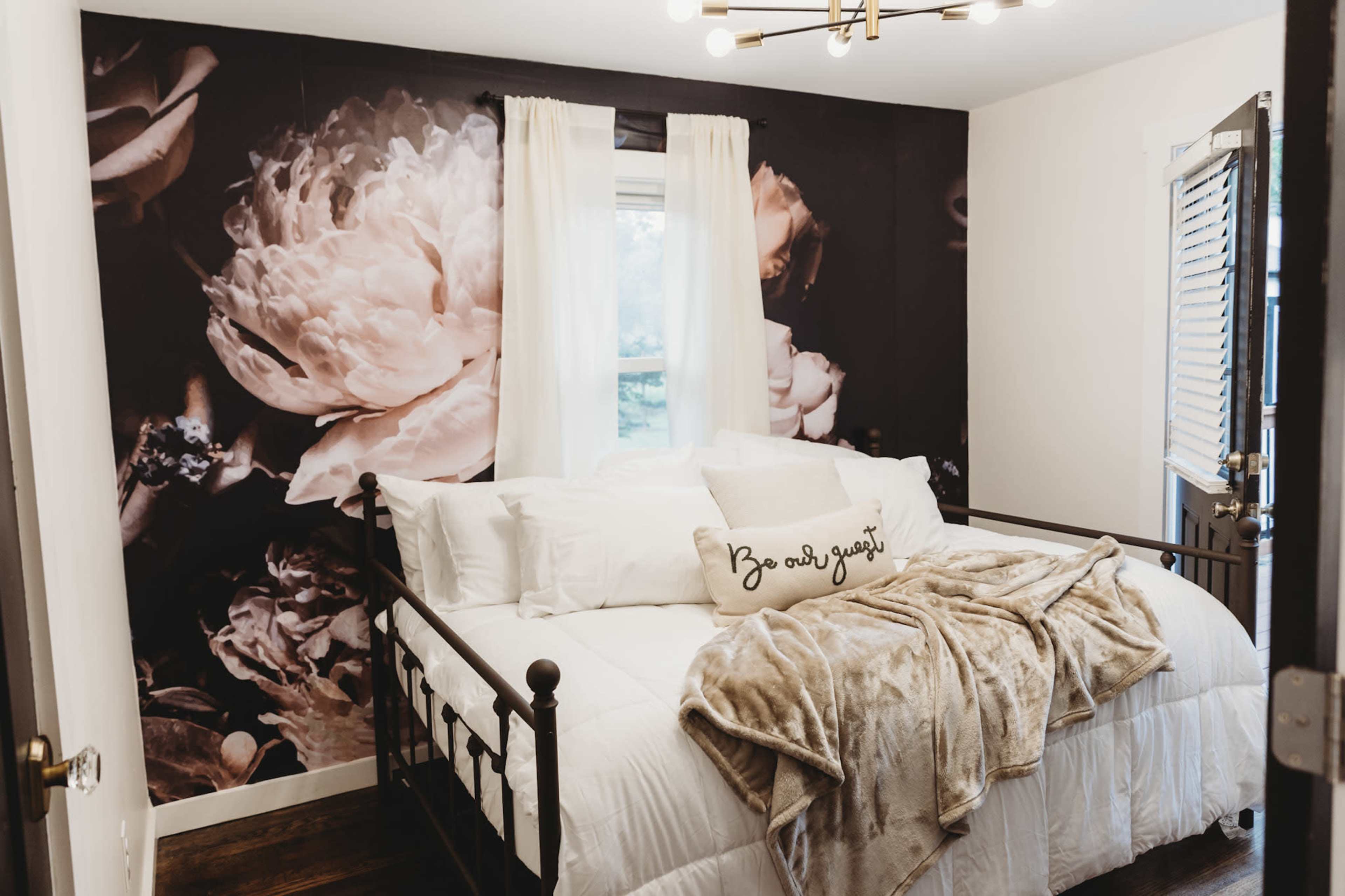 A bedroom with a floral accent wall, a wrought iron bed covered in white bedding, and a cozy throw blanket draped over it.