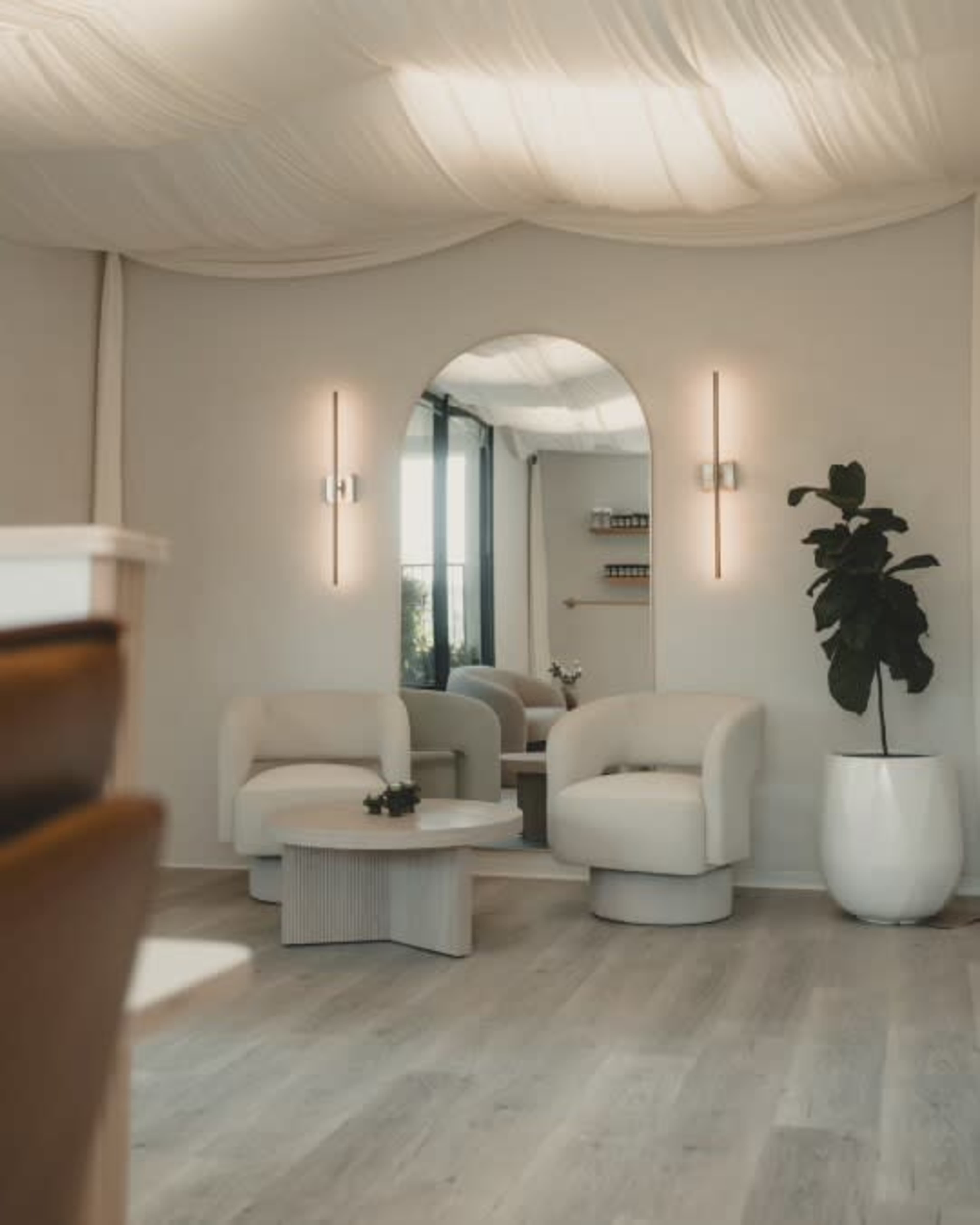 The image features a modern interior space with two white armchairs, a round coffee table, a tall plant in a white planter, and a wall-mounted mirror reflecting soft lighting.