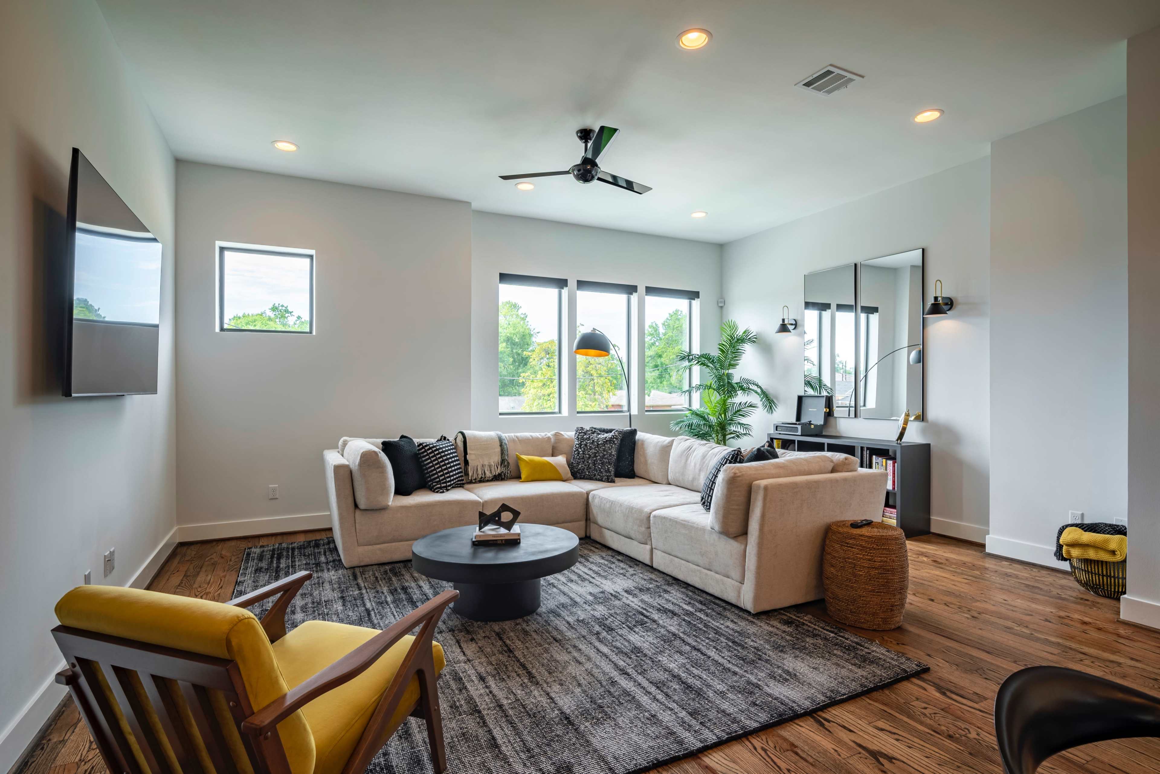 A modern living room features a sectional sofa, a round coffee table, and large windows that provide natural light.