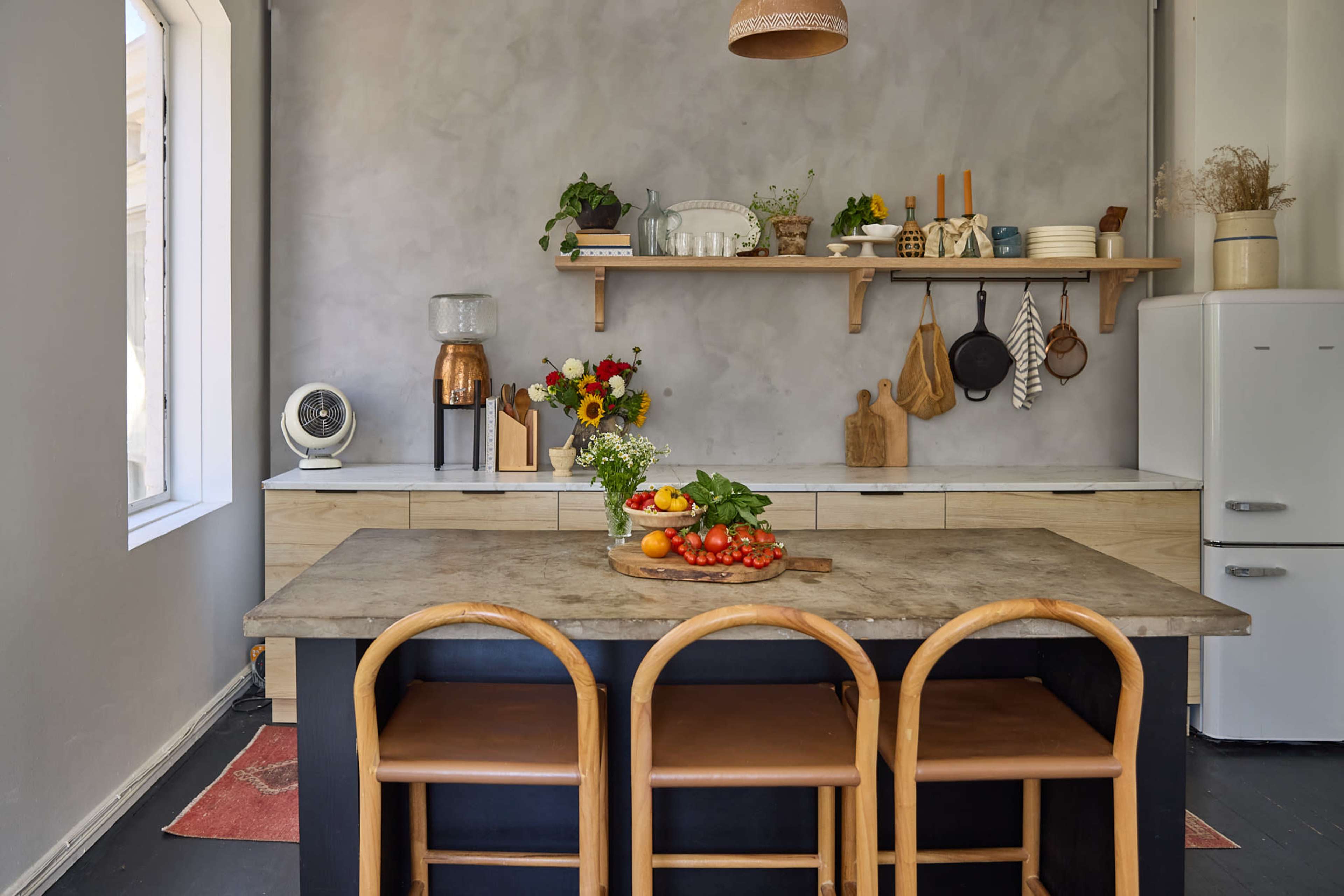 The kitchen features a wooden island with three chairs, a marble countertop, and an arrangement of fresh produce and flowers, alongside open shelving and a vintage refrigerator.