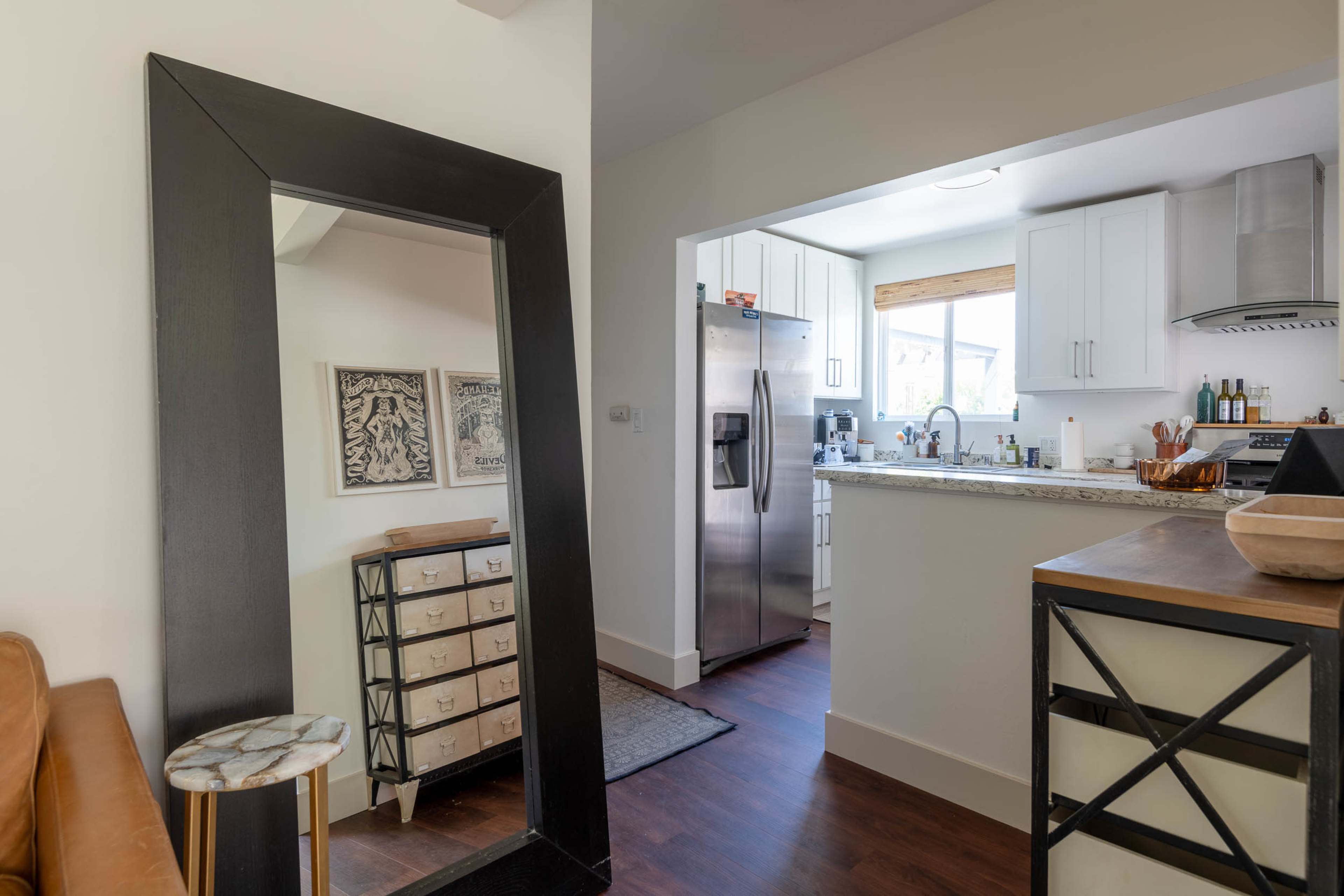 A modern interior features a wooden table, a large black mirror, and an open kitchen with white cabinetry and stainless steel appliances.