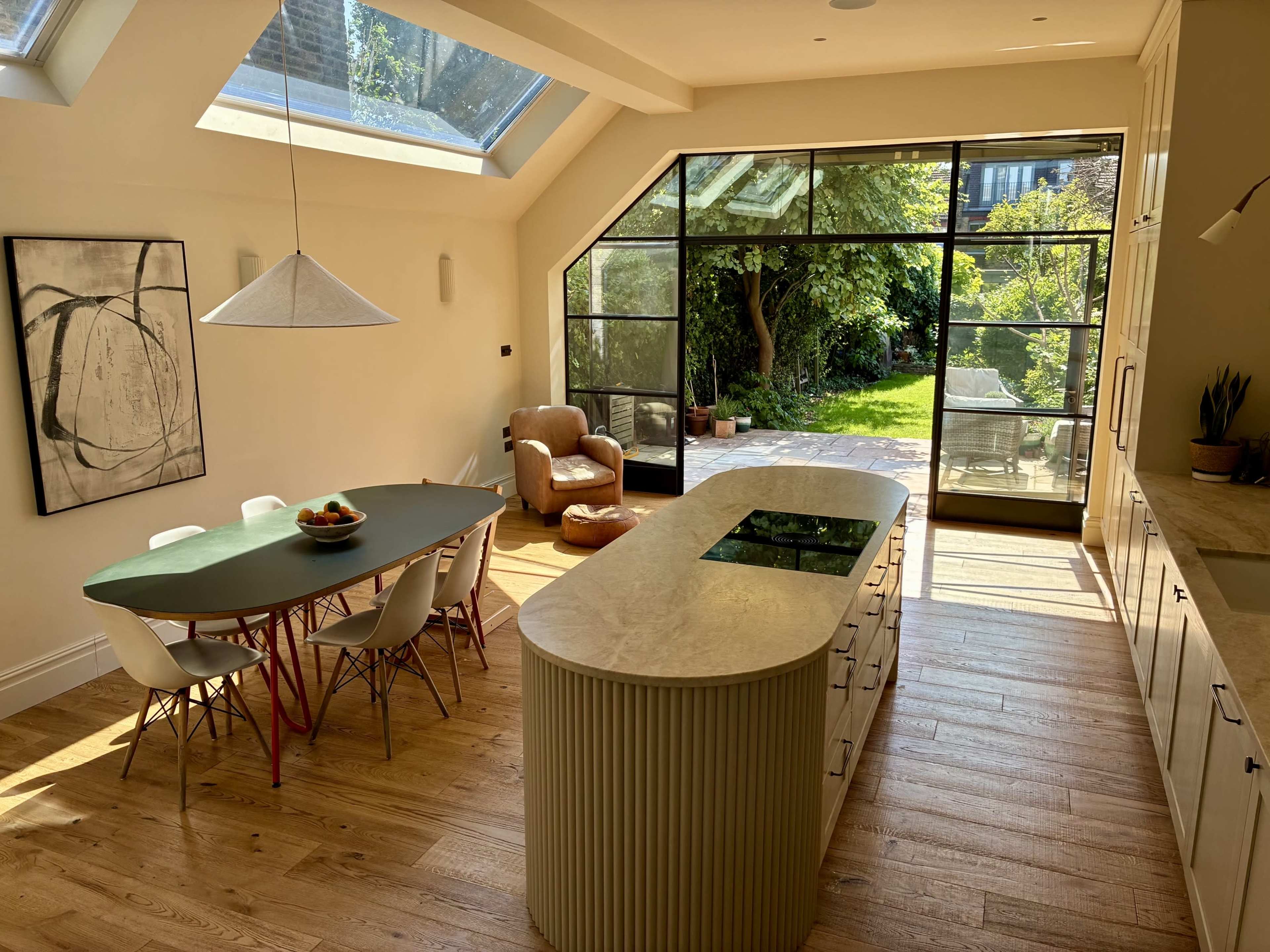 The image shows a modern kitchen and dining area with a large glass wall that opens to a garden, featuring a central kitchen island and a dining table surrounded by chairs.