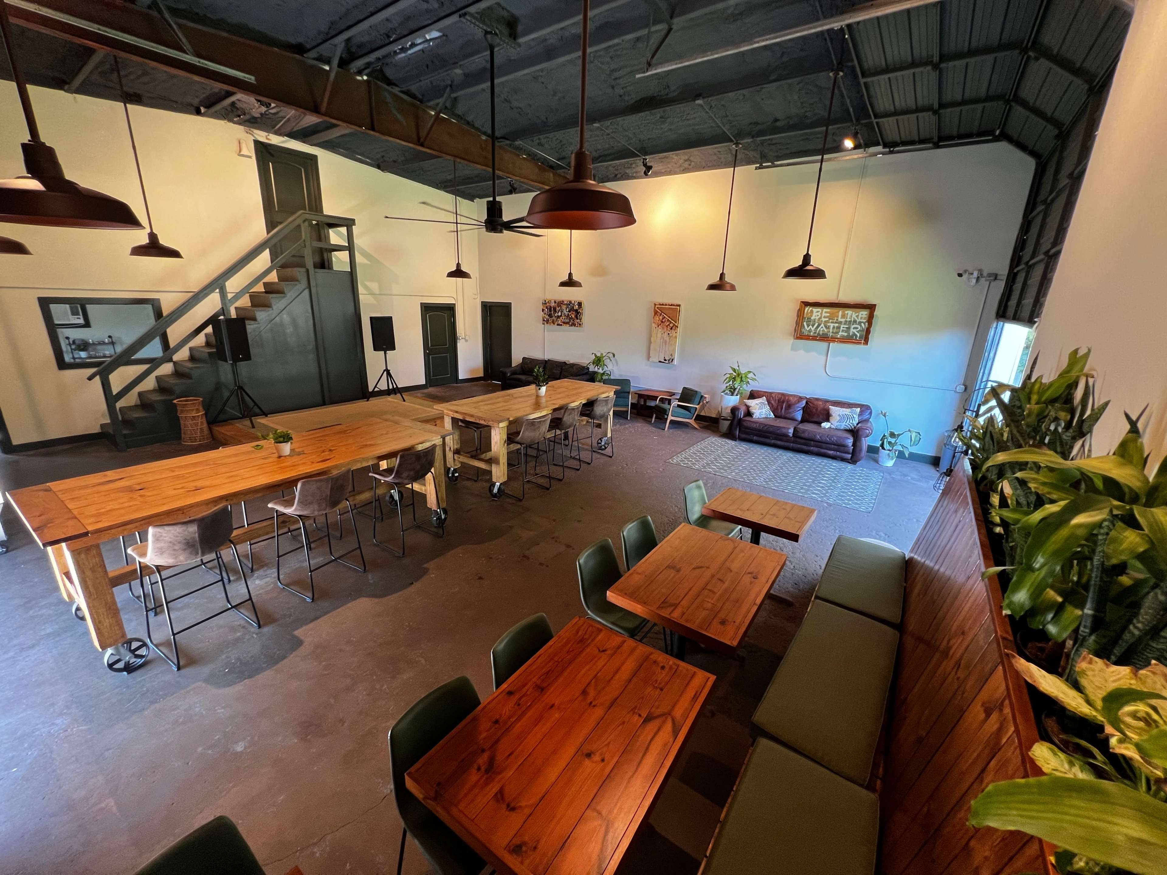 The image shows a spacious room with wooden dining tables, chairs, a sofa area, and a staircase, adorned with plants and decorative artwork.