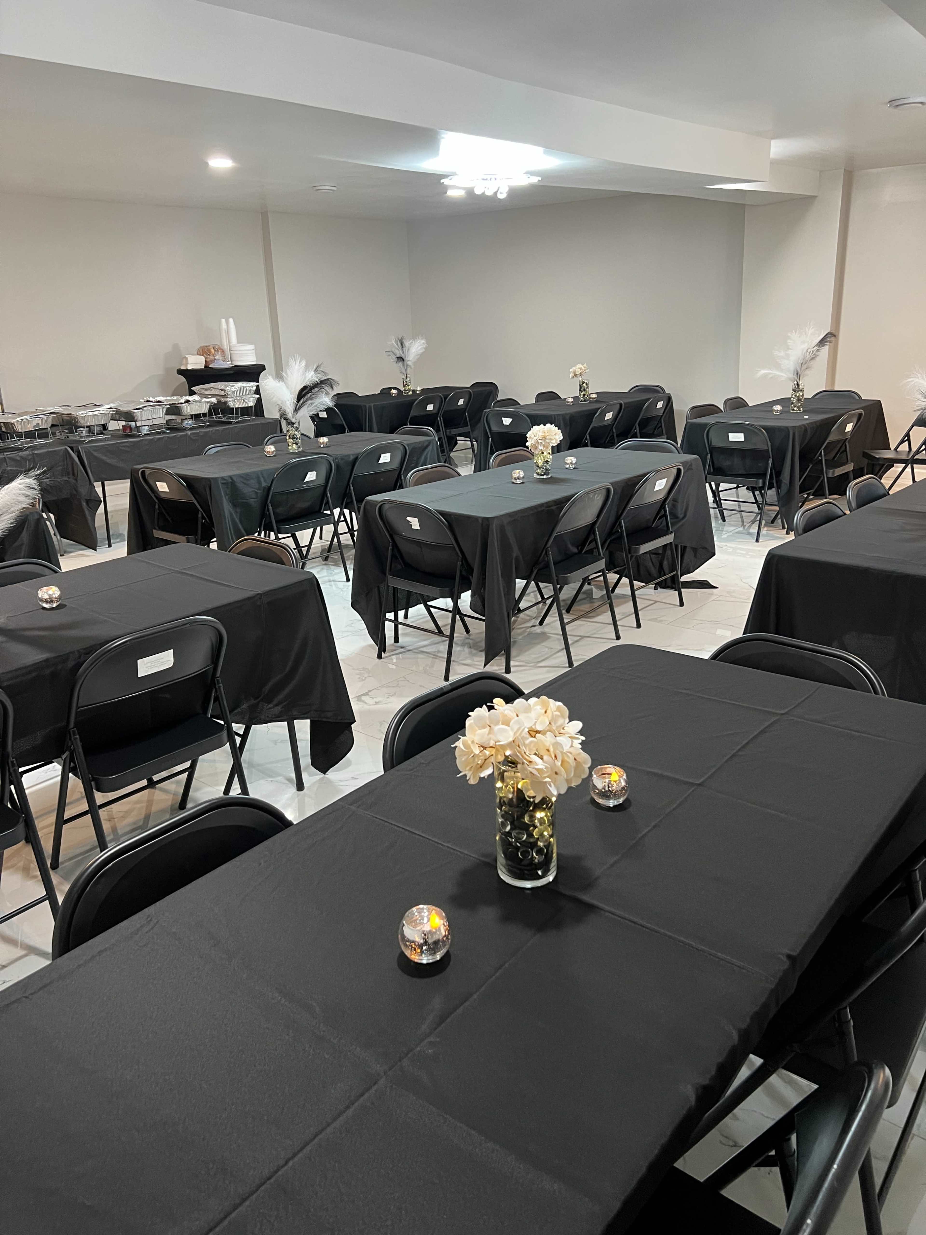 The image shows a banquet hall set with multiple black-covered tables, each adorned with small floral centerpieces and candles.