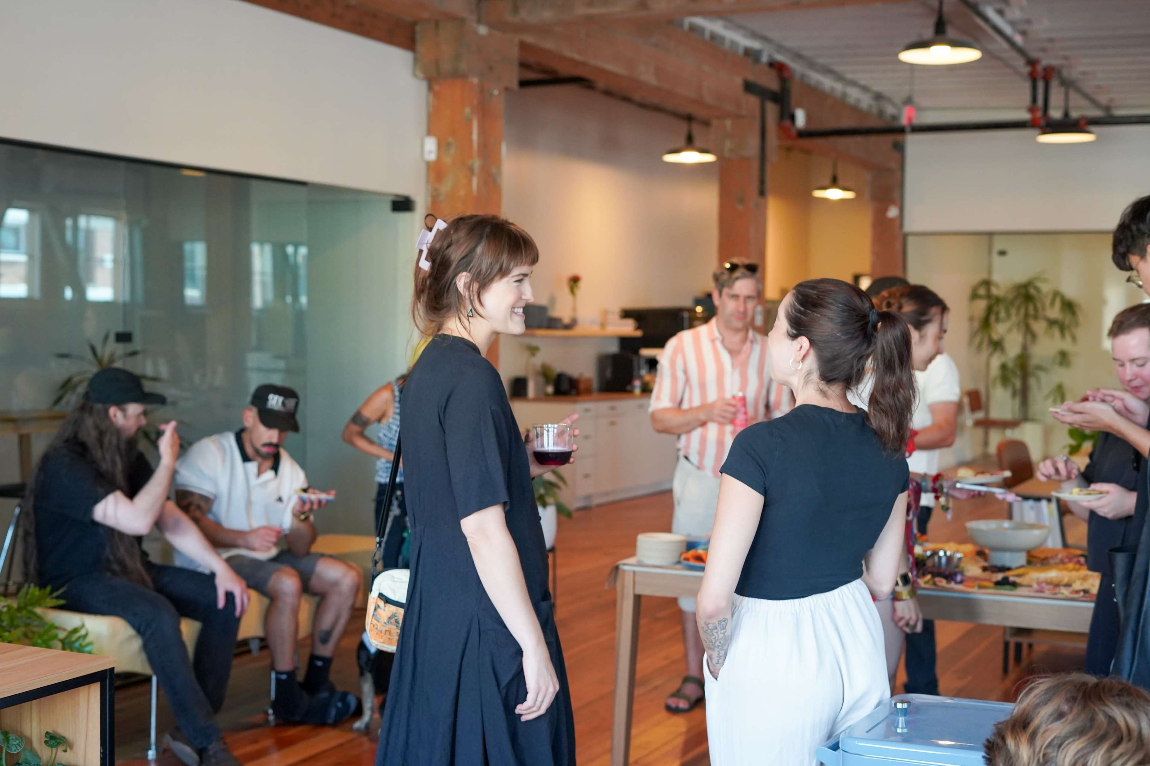 A group of people socializes in a modern office space with wooden beams, enjoying food and drinks during a gathering.