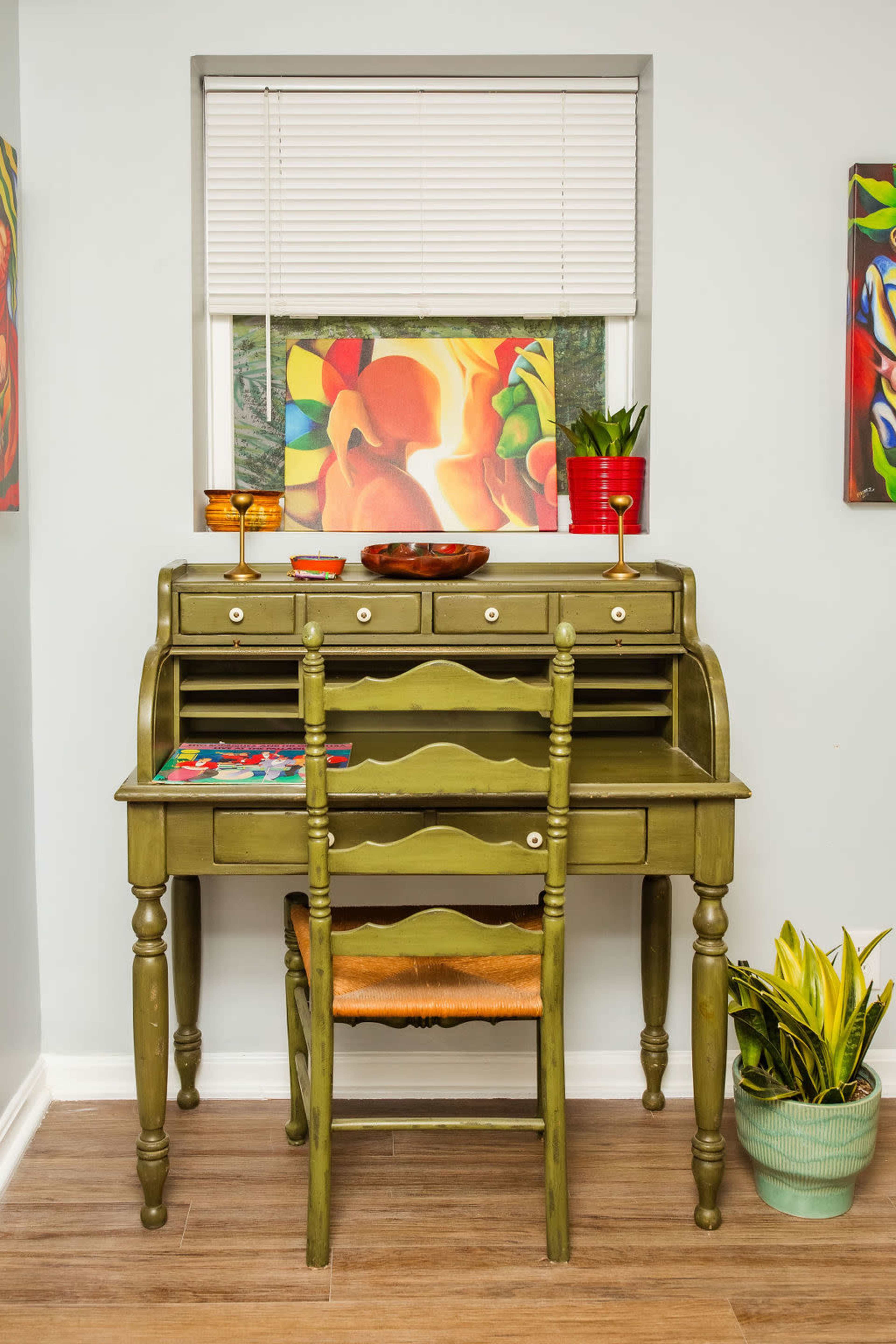 A green writing desk with a single chair is positioned against a wall under a window, adorned with artwork and a small potted plant.