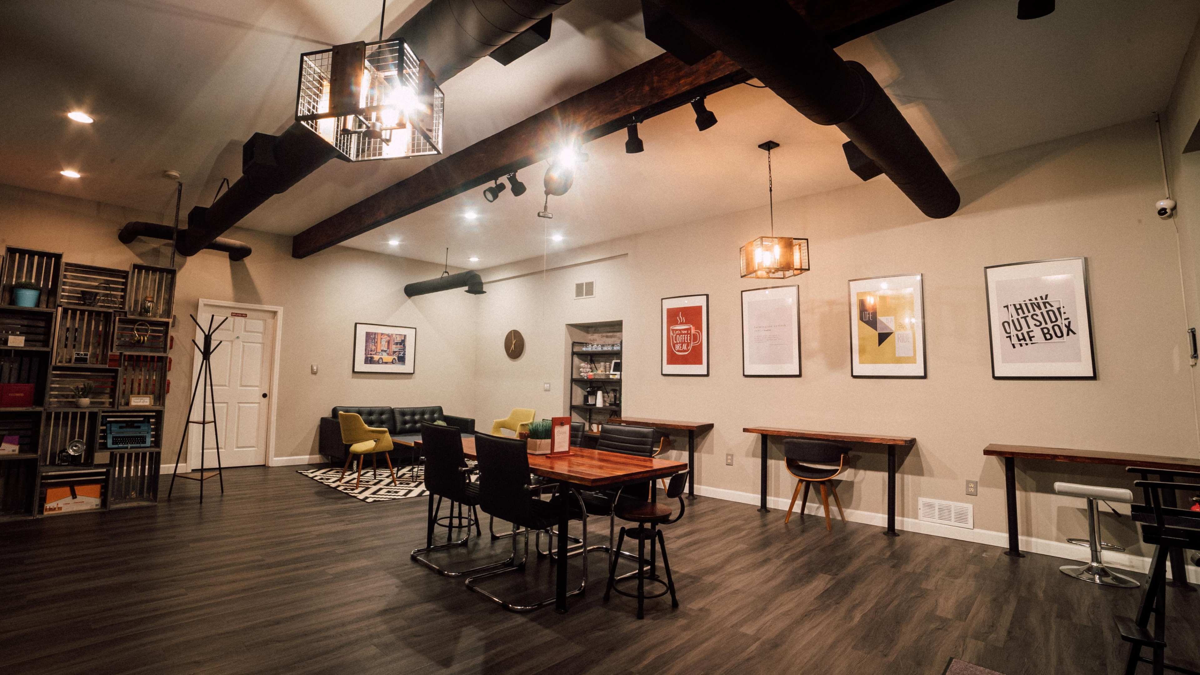 The image shows a modern, open room with wooden beams, a dining table, seating area, and framed artwork on the walls.