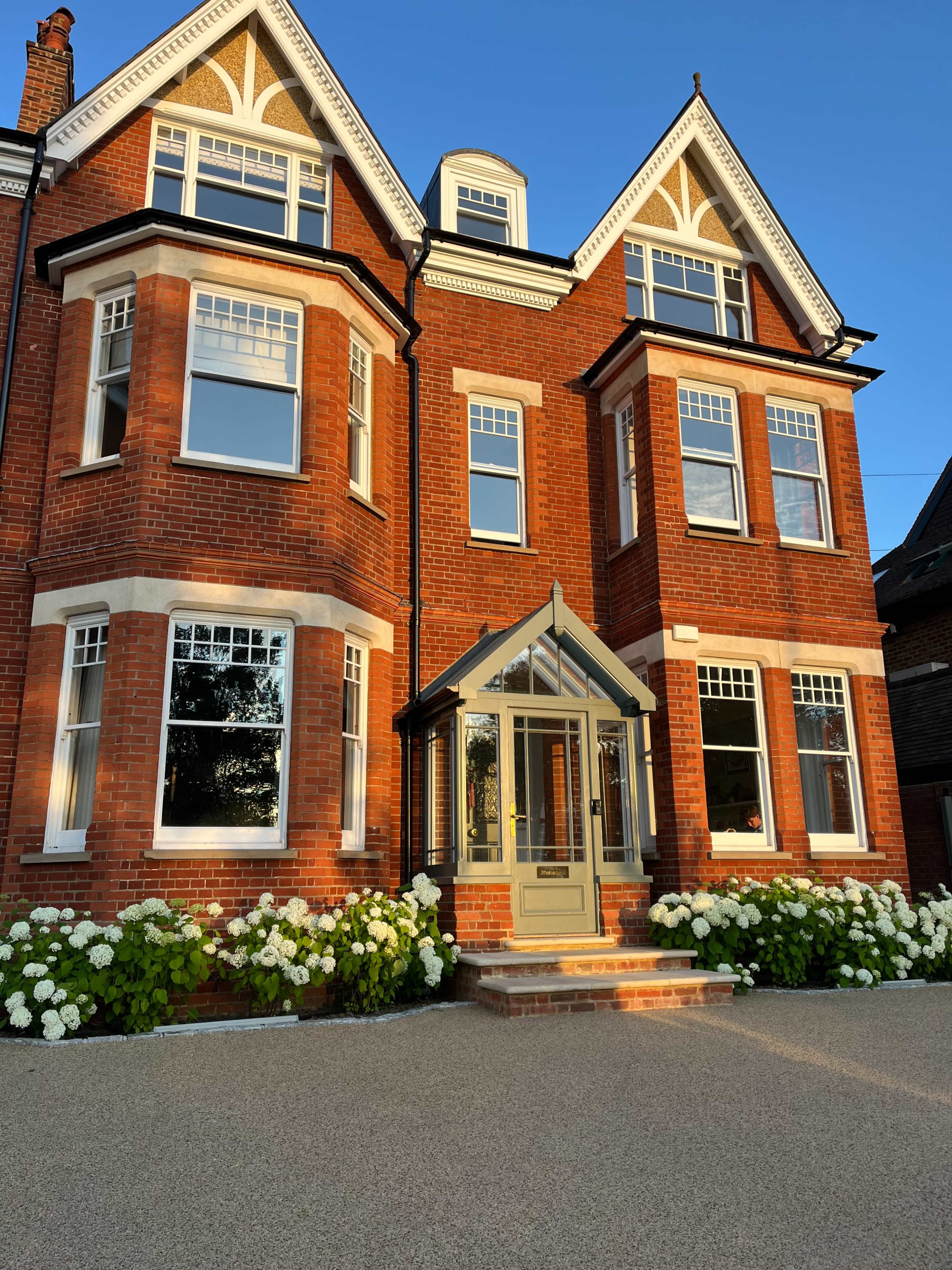 A red brick house with large windows, a glass front porch, and a flower garden of white hydrangeas surrounds the entrance.