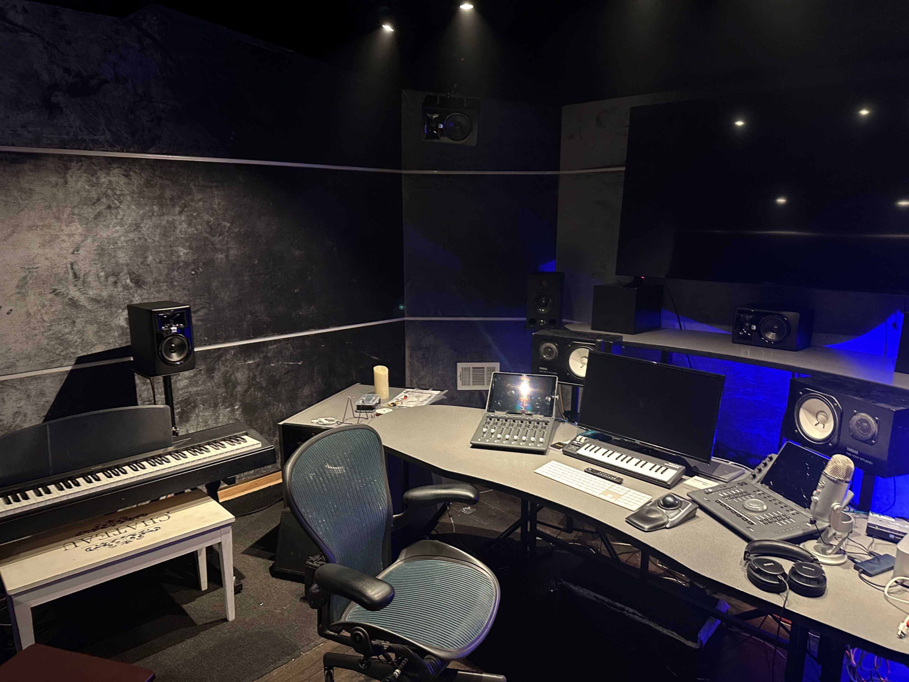 The image shows a music production studio with a desk equipped with a computer, audio mixer, and speakers, alongside a keyboard and a microphone.