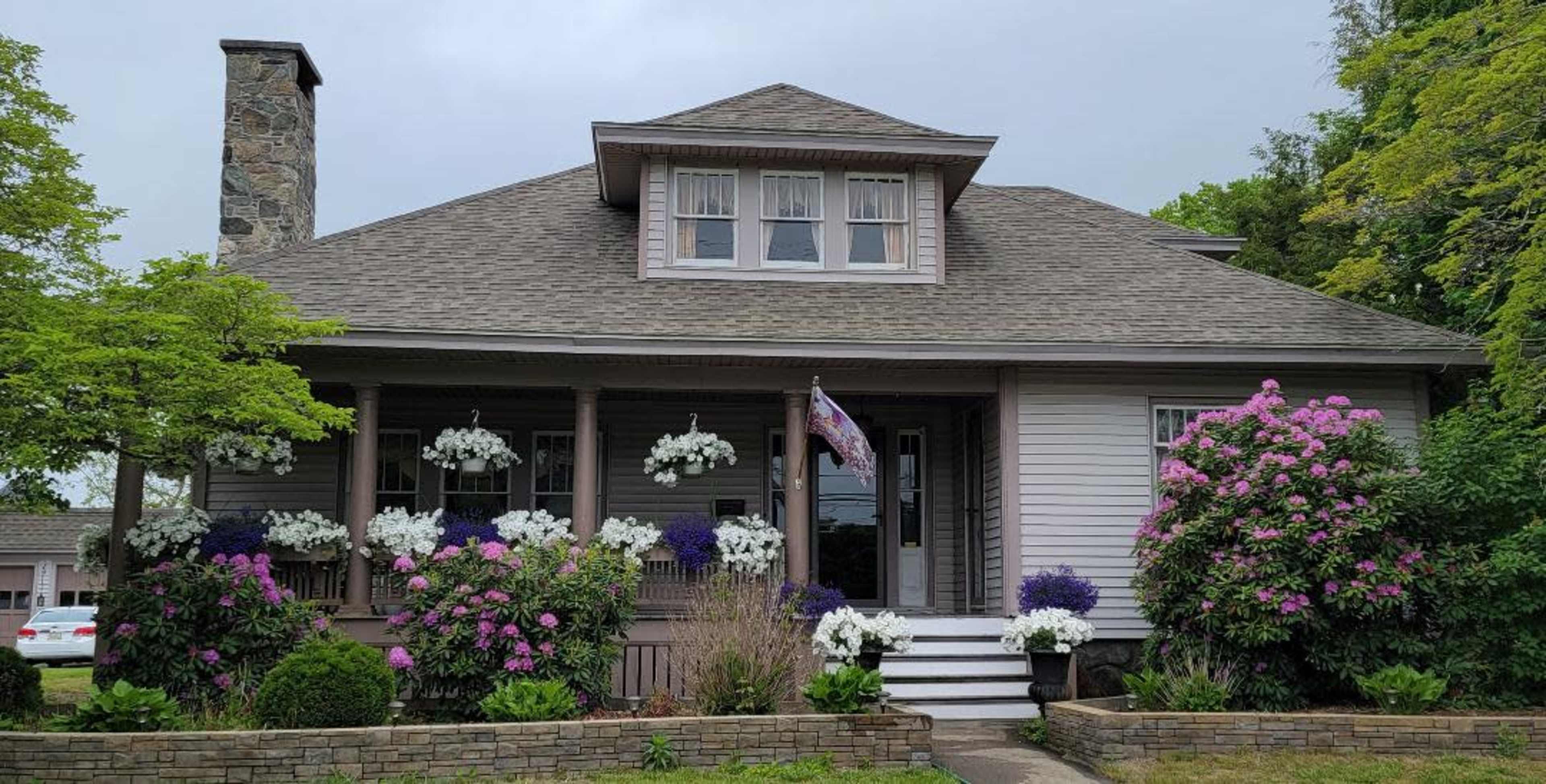 A two-story house with a stone column features a welcoming porch adorned with hanging flower baskets and colorful flower beds.