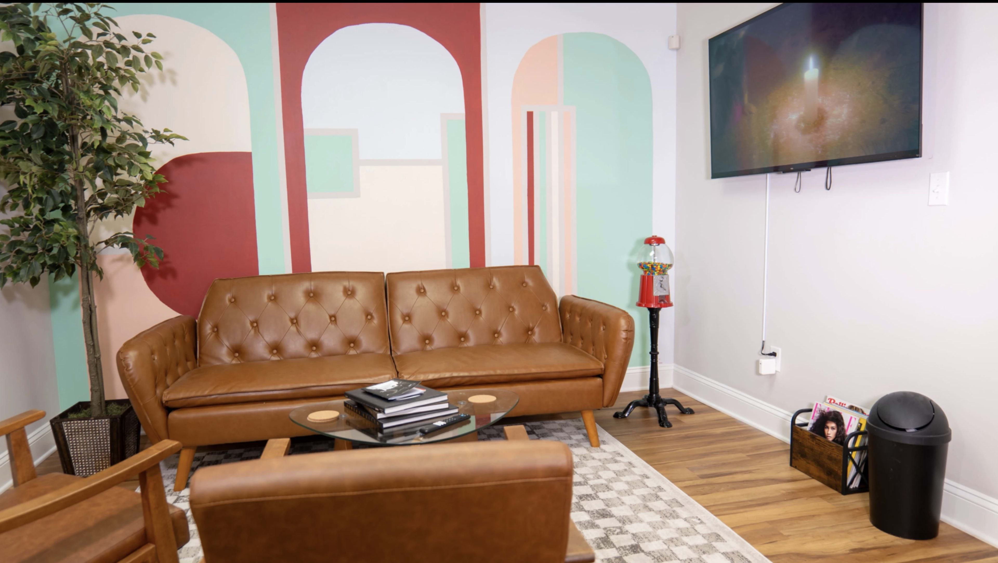 The scene depicts a modern living room with a brown leather couch, a coffee table stacked with magazines, a decorative wall with colorful geometric shapes, and a flat-screen TV mounted on the wall.