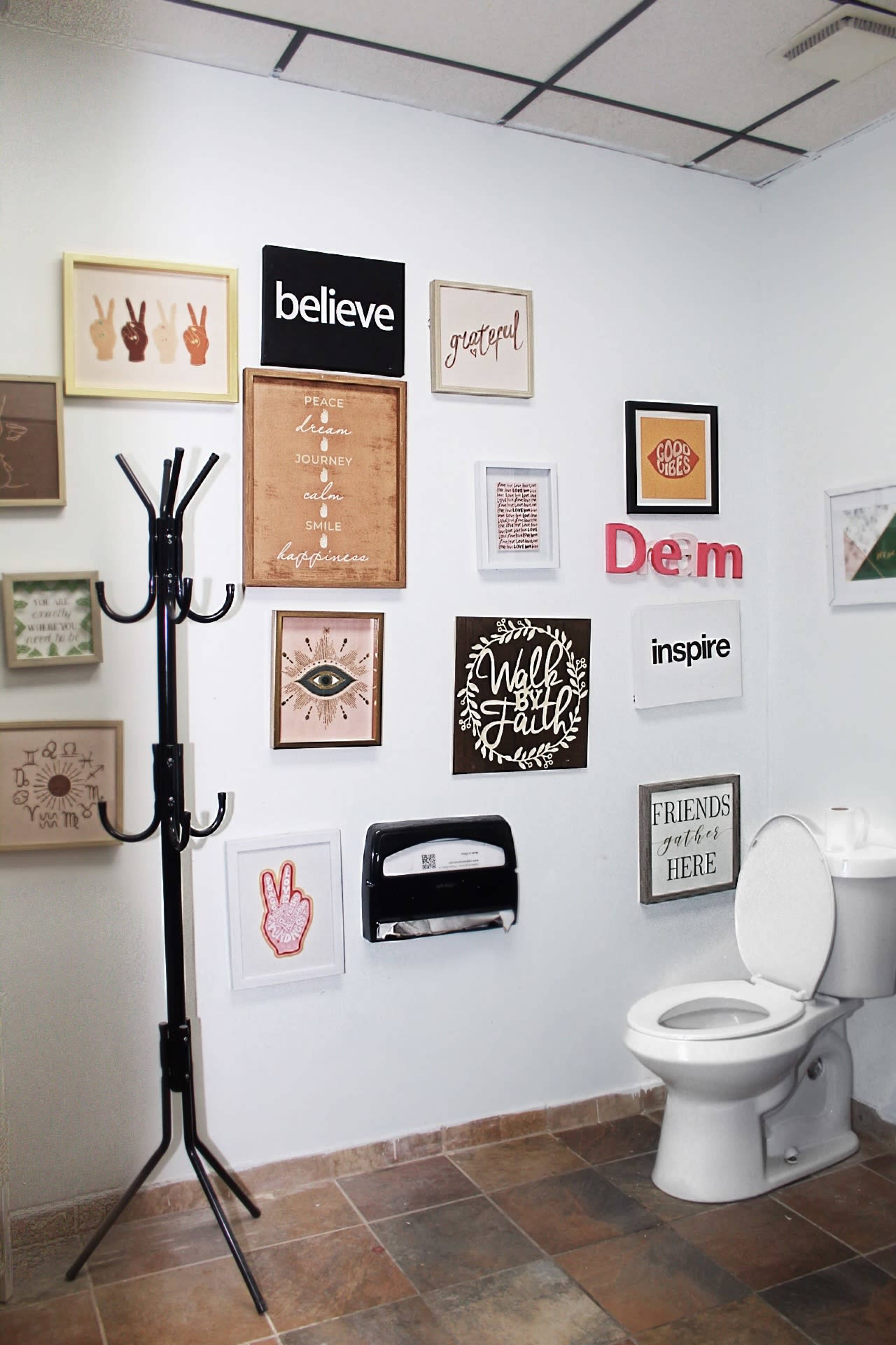 A bathroom features a white wall adorned with various framed motivational quotes and a black coat rack next to a toilet and paper dispenser.