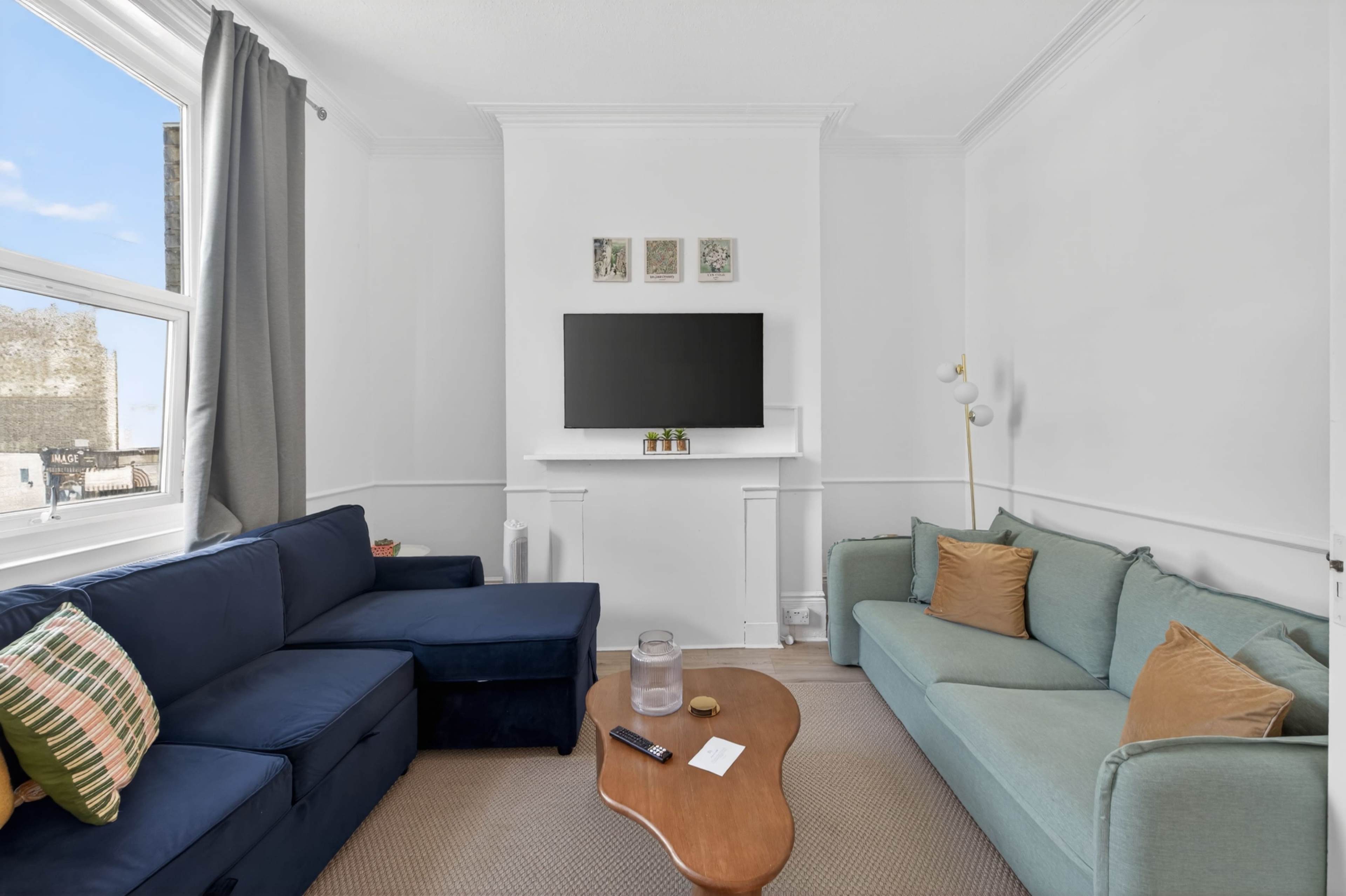 The image shows a modern living room with two sofas, a wooden coffee table, a TV mounted on the wall, and large windows.