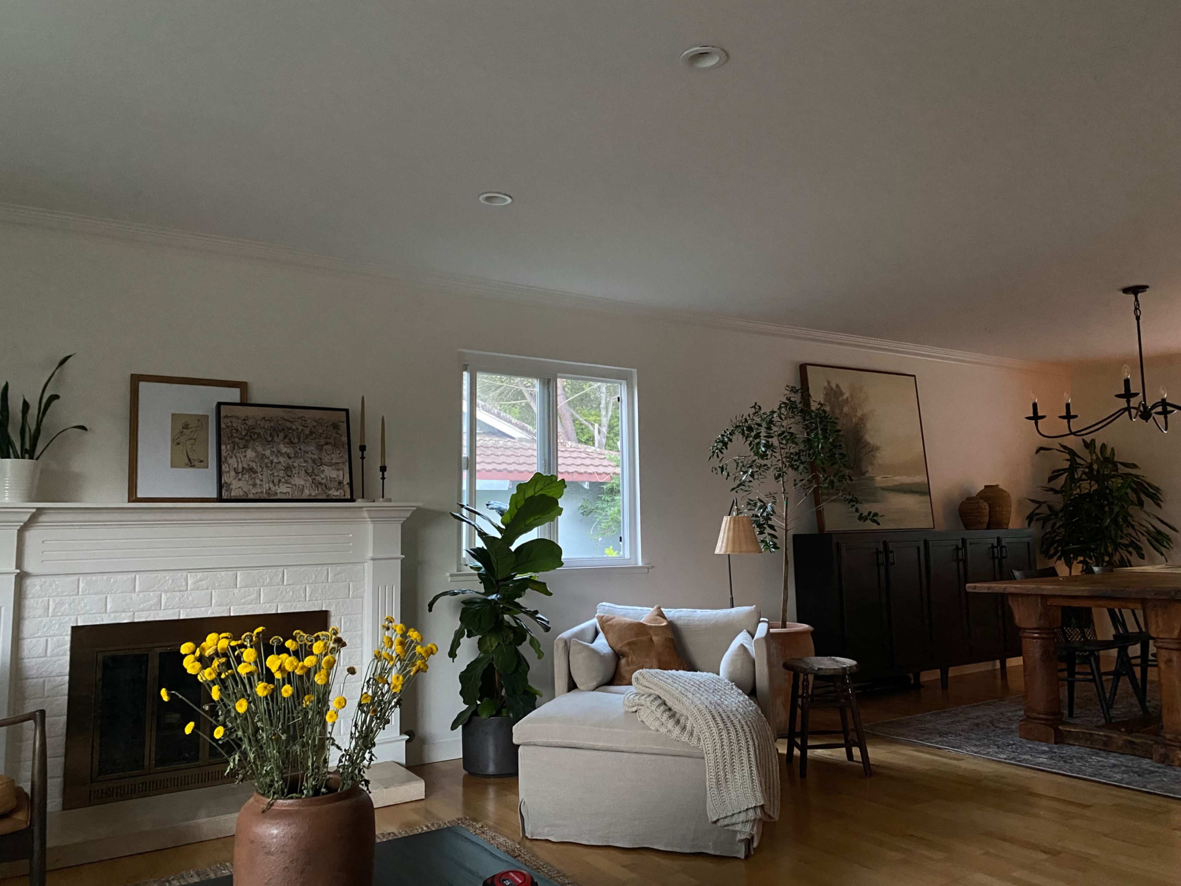 A cozy living room featuring a large window, a white-brick fireplace, and a mix of plants and furniture, including a sectional sofa, dining table, and side chair.