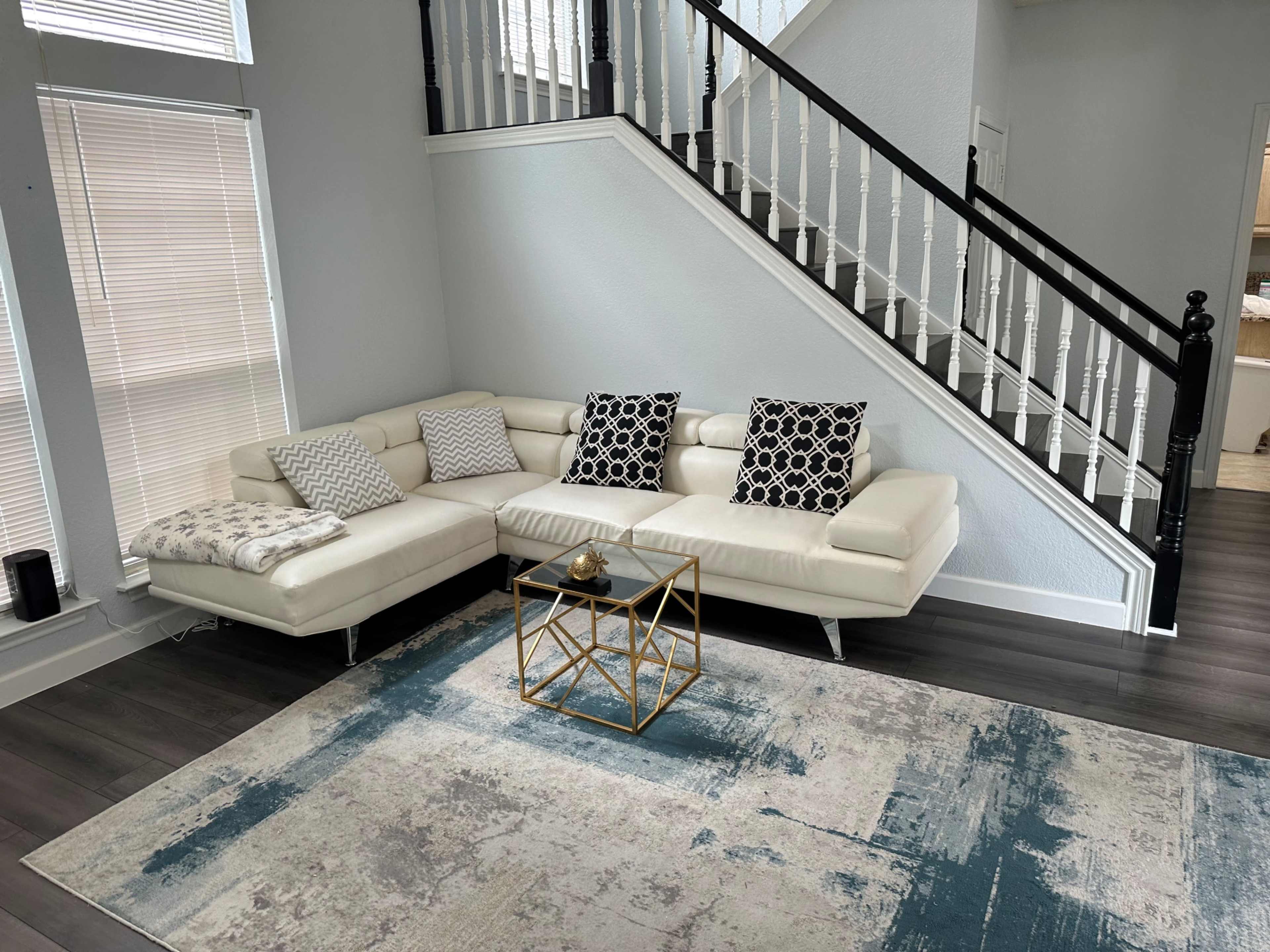 A modern living room featuring a white sectional sofa with patterned pillows, a gold geometric coffee table, and a textured area rug, set against a backdrop of a staircase with black railing.