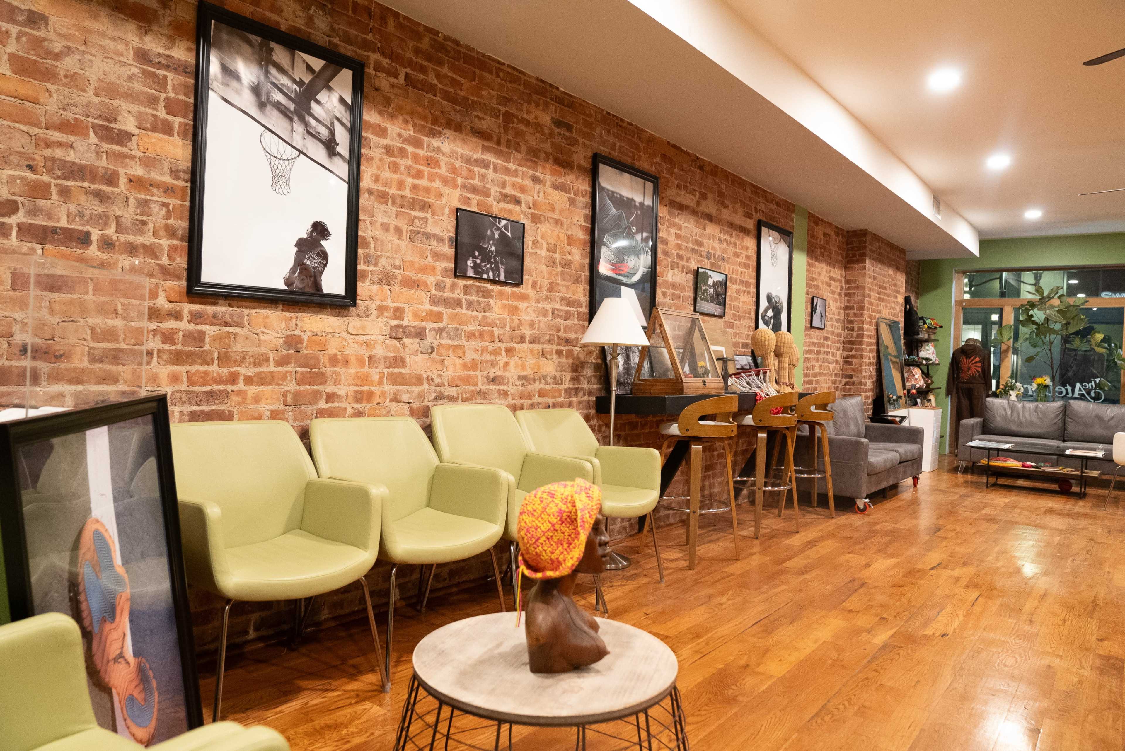 The image shows a warm-toned interior of a room featuring exposed brick walls, green chairs, framed photographs, and a wooden floor.