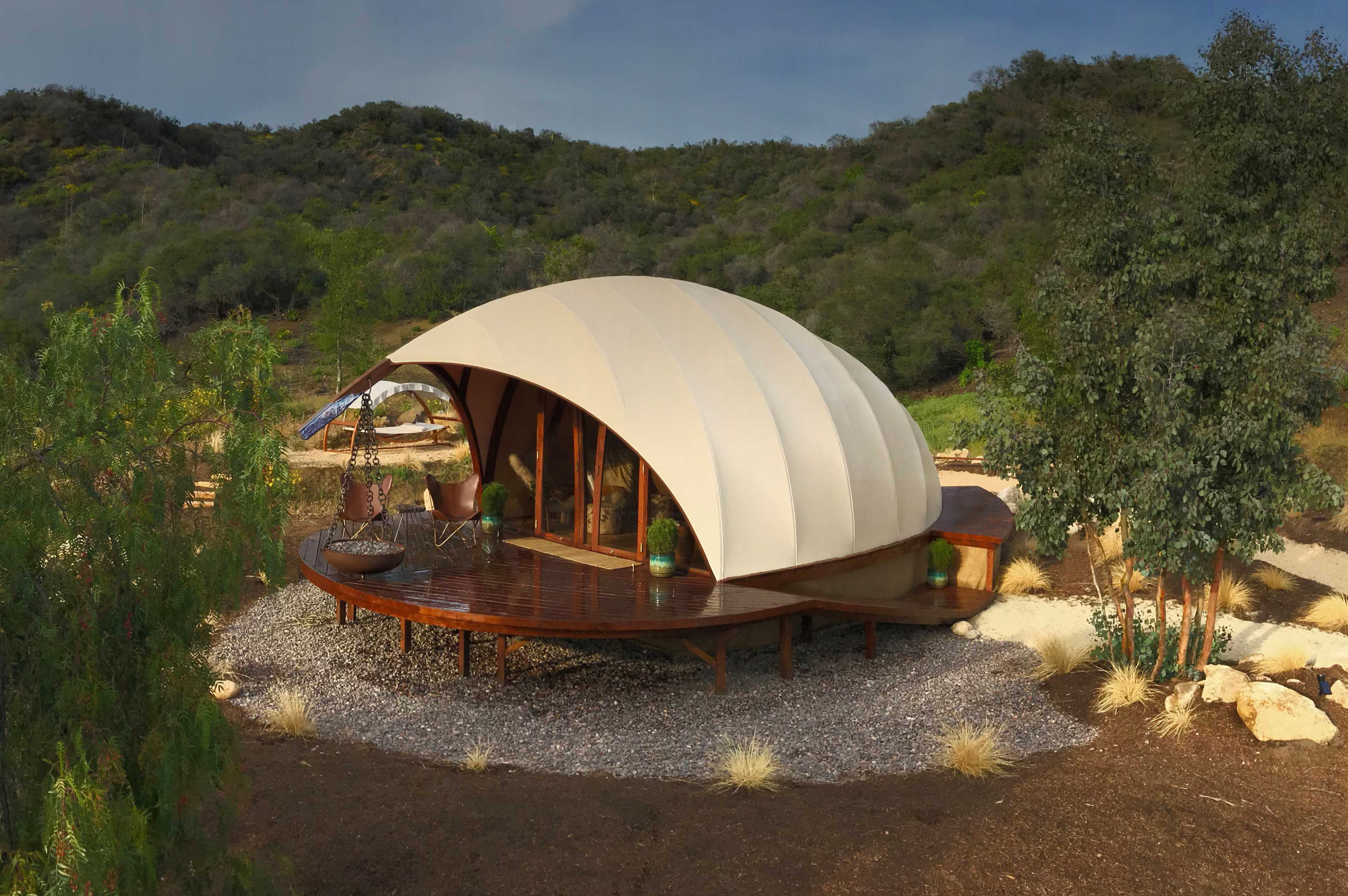A uniquely designed, curved, fabric-covered structure sits on a wooden platform surrounded by gravel and sparse vegetation.