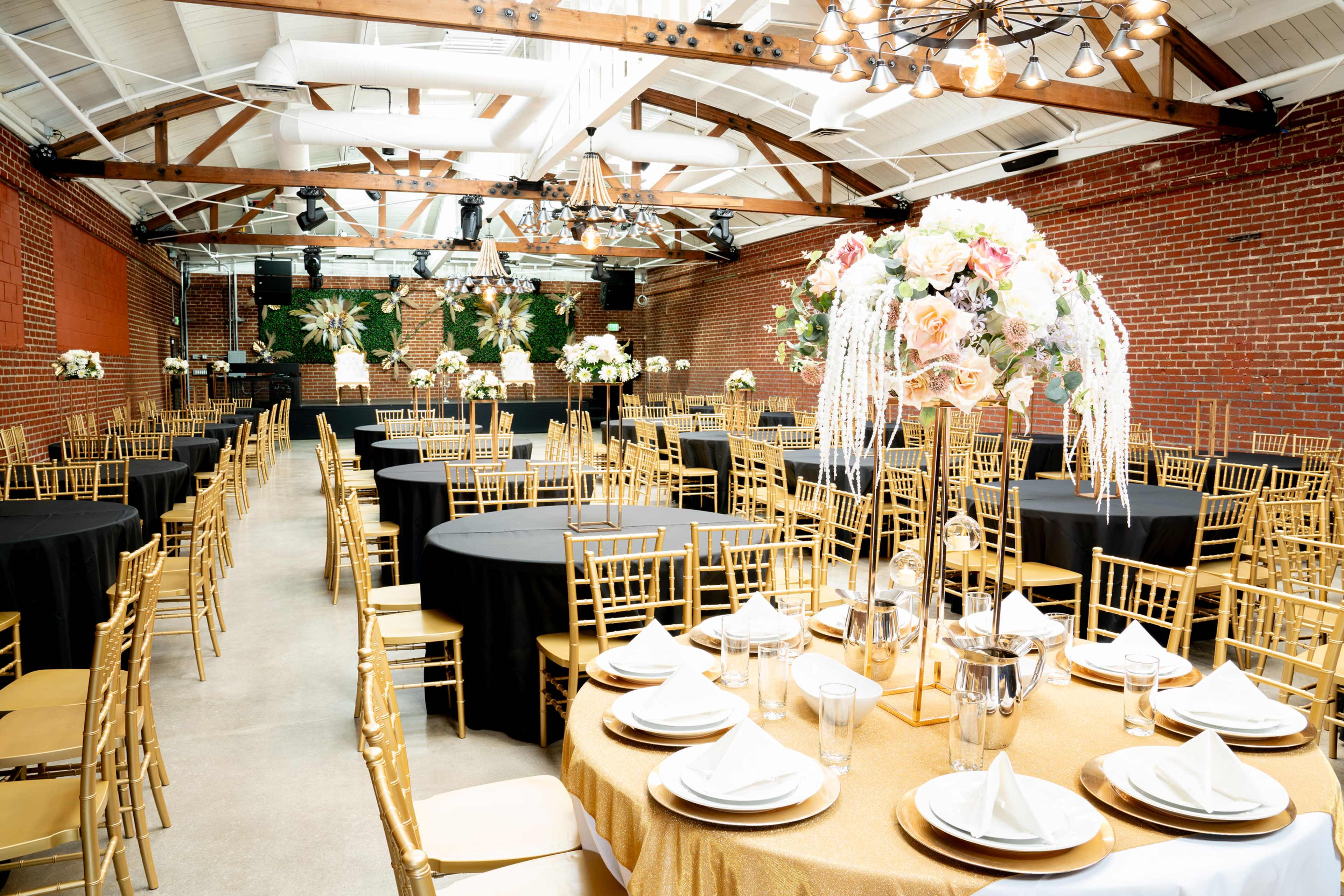 A spacious event venue features round tables set with elegant tableware and decorative floral centerpieces, surrounded by gold chiavari chairs.
