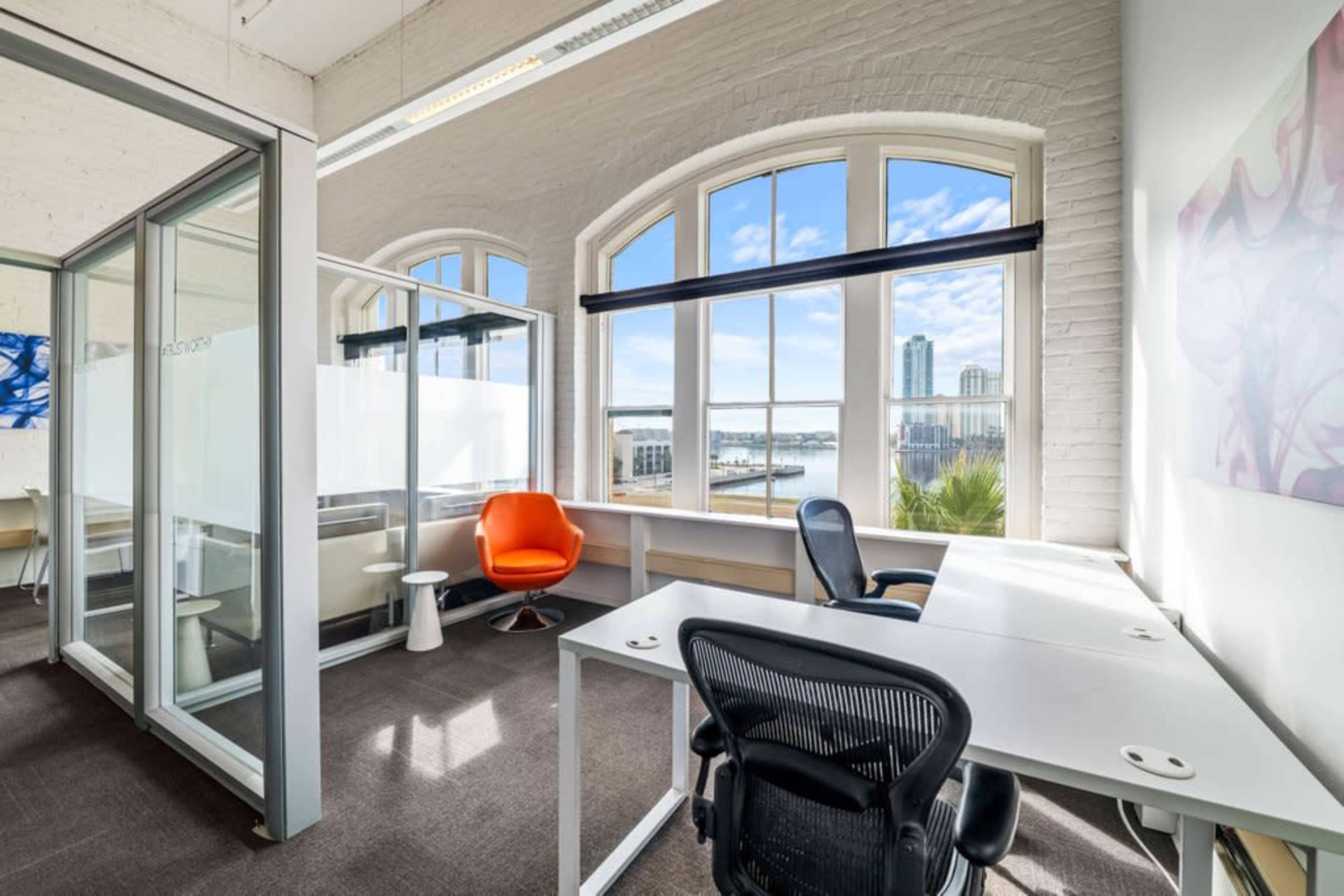 The image shows a modern office space featuring large windows with a view of a waterfront and city skyline, along with a bright orange chair and white desks.
