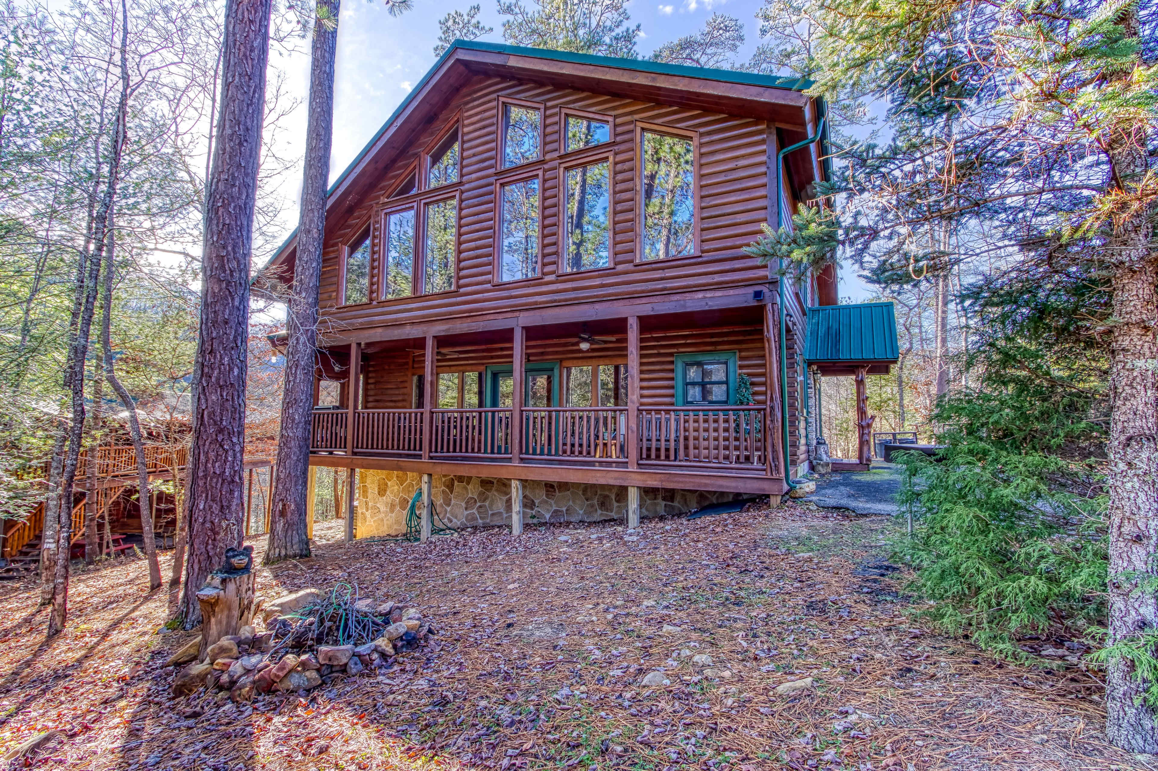A large log cabin with multiple windows and a covered porch is situated among trees in a wooded area.