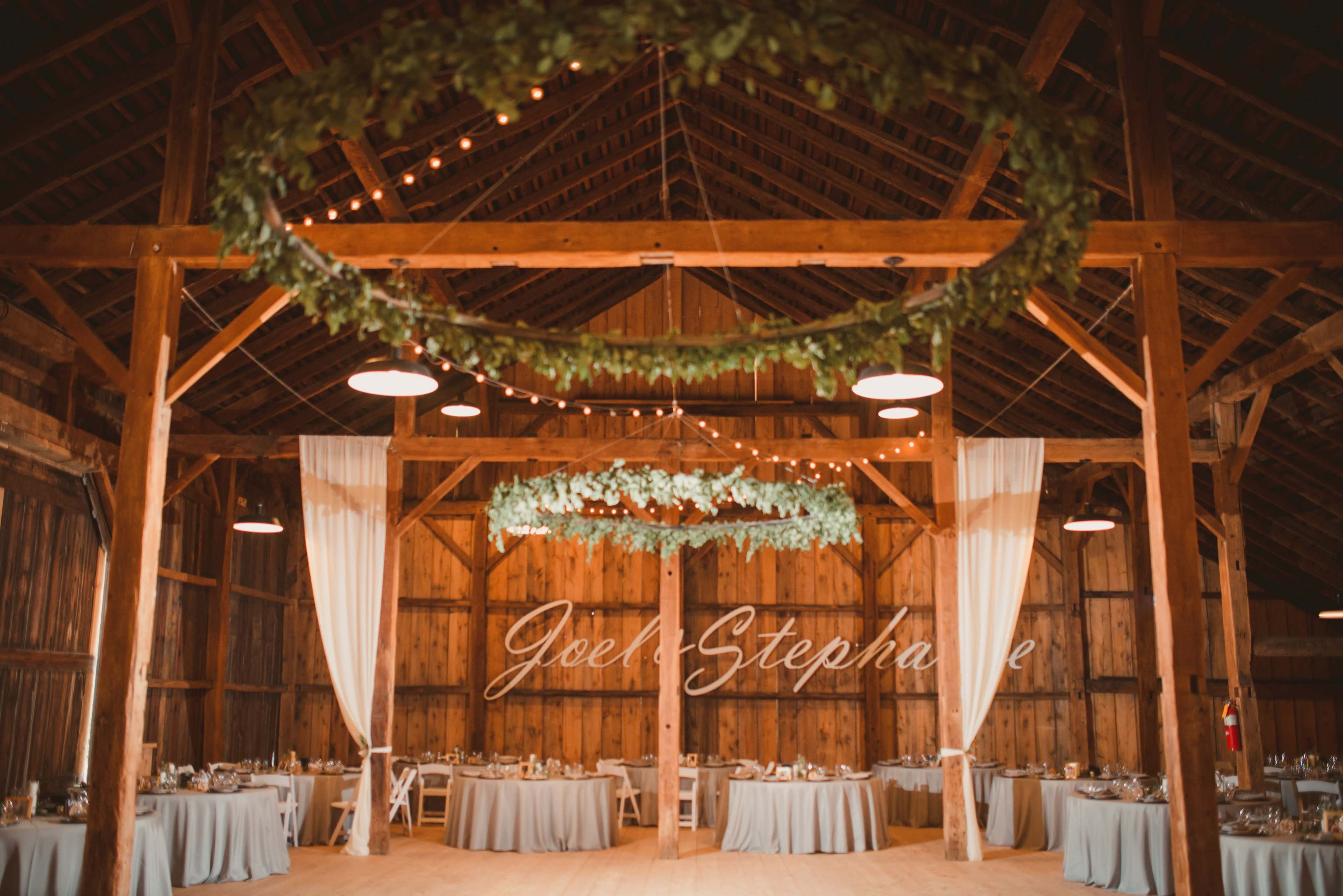 The interior of a rustic barn is decorated for an event, with round tables set and greenery wreaths hanging from the beams above.