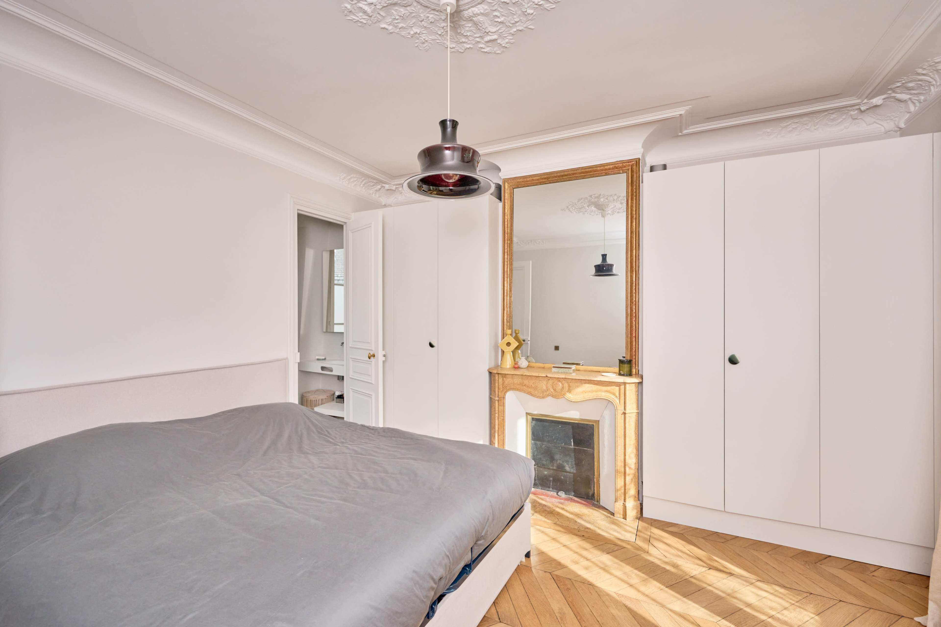 The image shows a bedroom with a bed, a mirrored dresser, and white cabinetry against a light-colored wall.